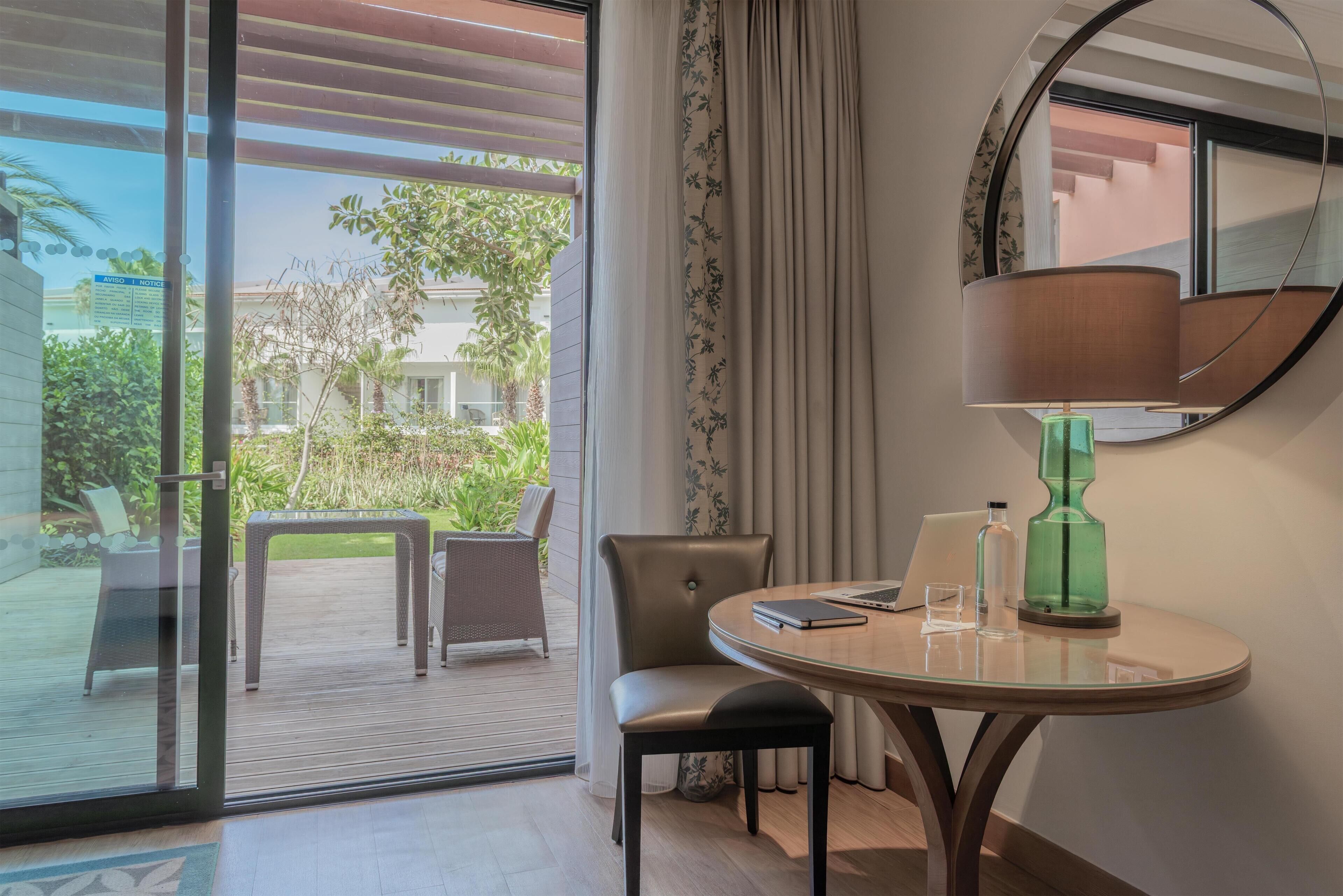 Une chambre d’hôtel avec une porte vitrée donnant sur une terrasse extérieure, une table ronde avec une lampe, une chaise et une petite table d’appoint.