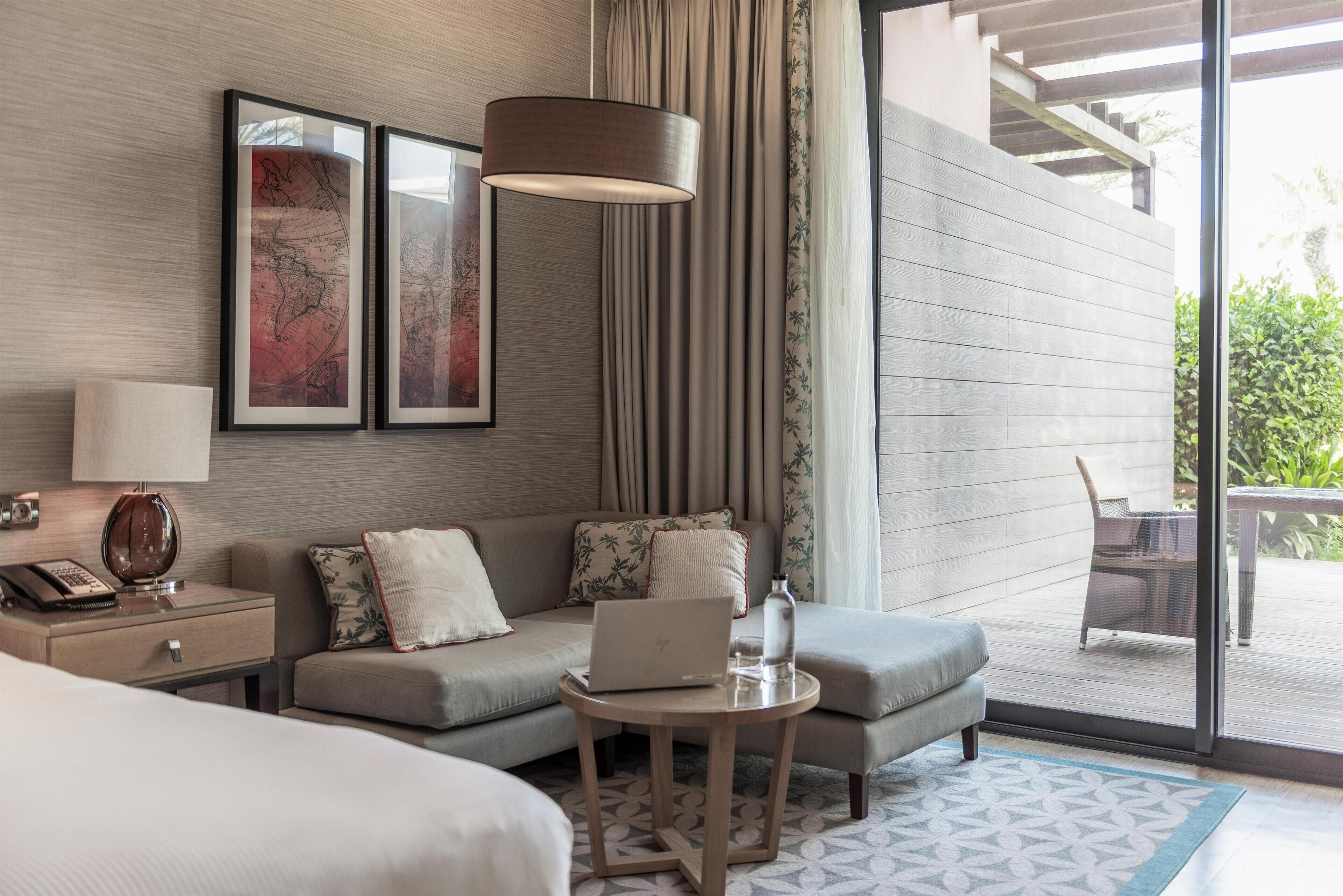 Ein Hotelzimmer mit einem Bett, einem Sofa, einem kleinen Tisch mit einem Laptop, einer Lampe, einem Telefon und einem Fenster mit Vorhängen.