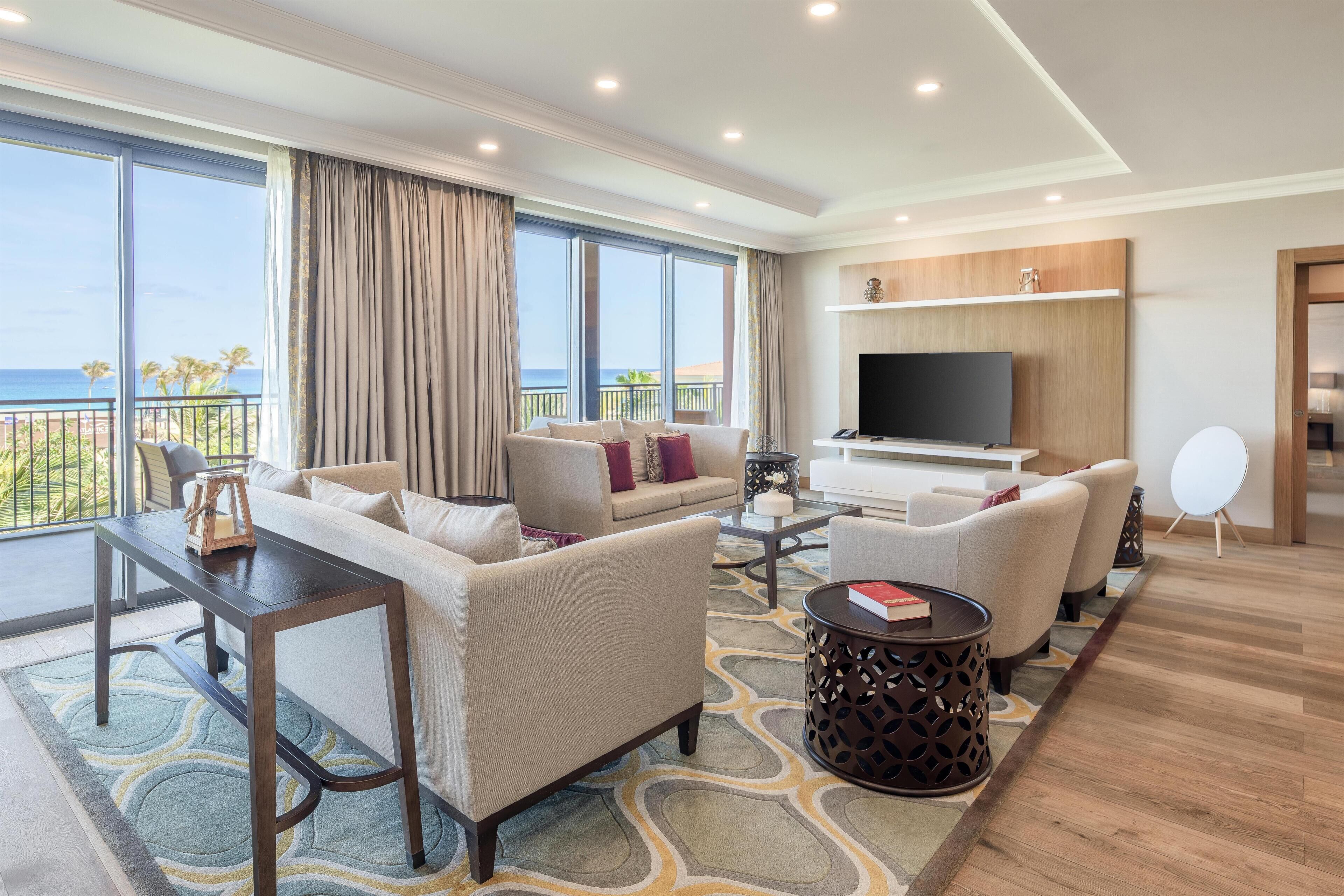 Une chambre d’hôtel moderne dotée d’un balcon, d’un ensemble de canapés, d’une table basse, d’un téléviseur et d’une petite table d’appoint.