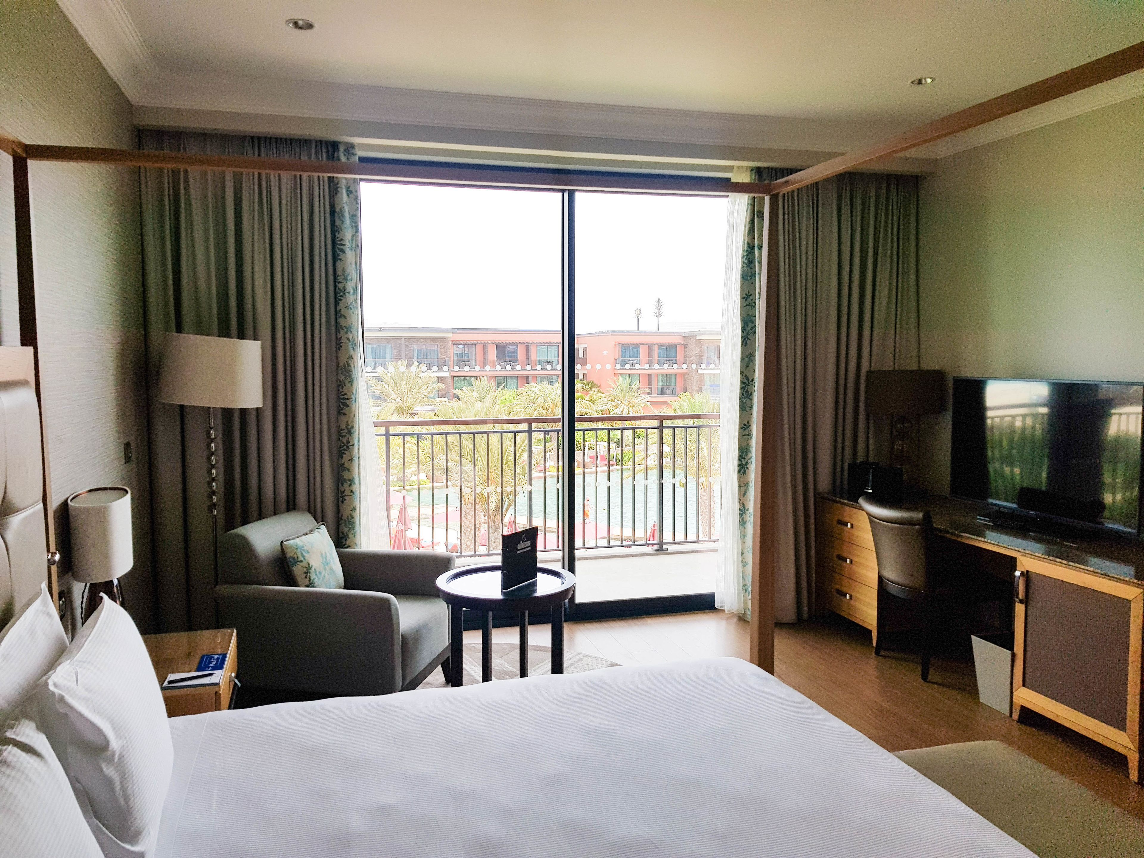 A hotel room with a bed, a desk, a chair, a TV, and a balcony with a view of buildings and a pool.