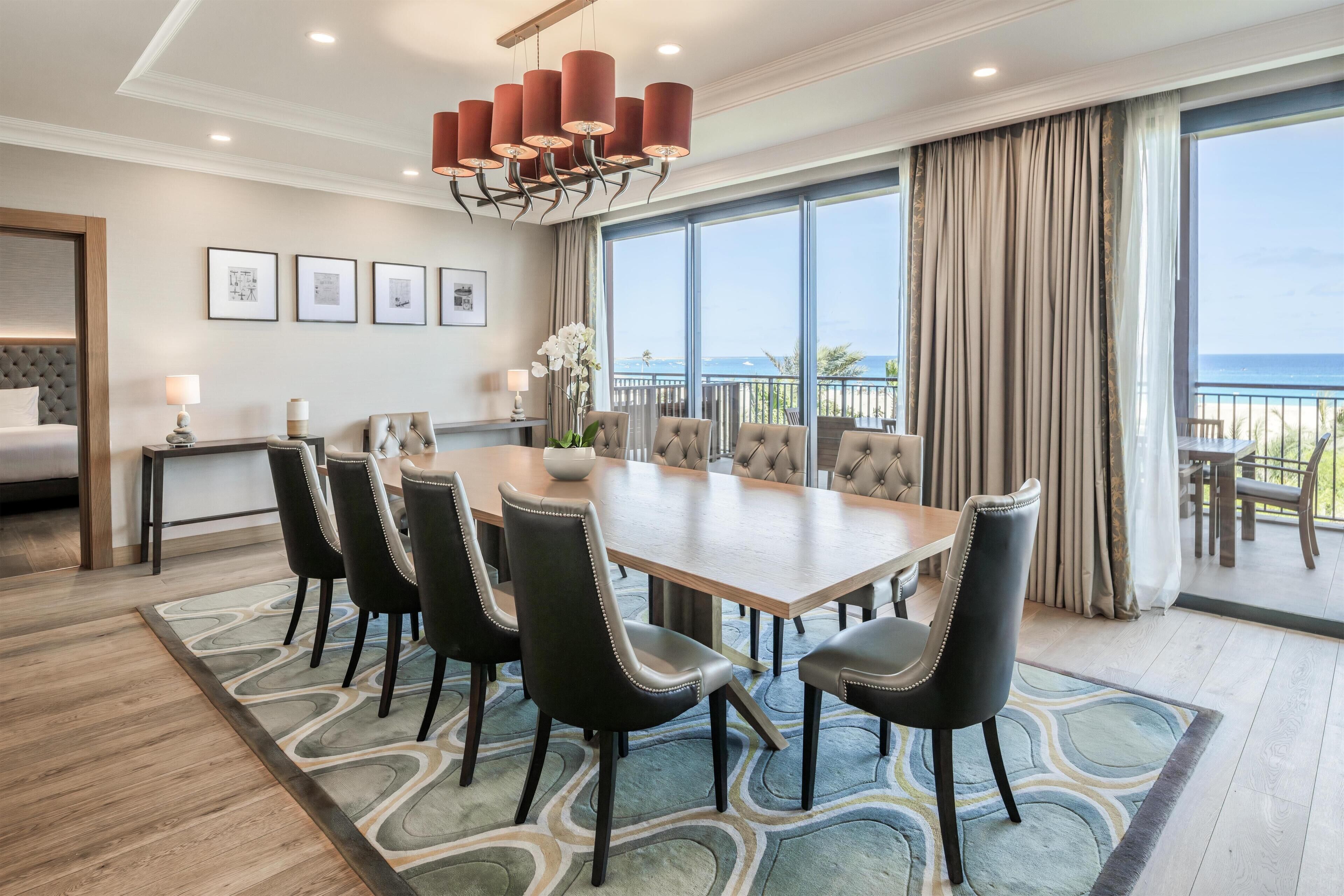 A hotel room with a large dining table, multiple chairs, a chandelier, a bed, and a balcony with a view.