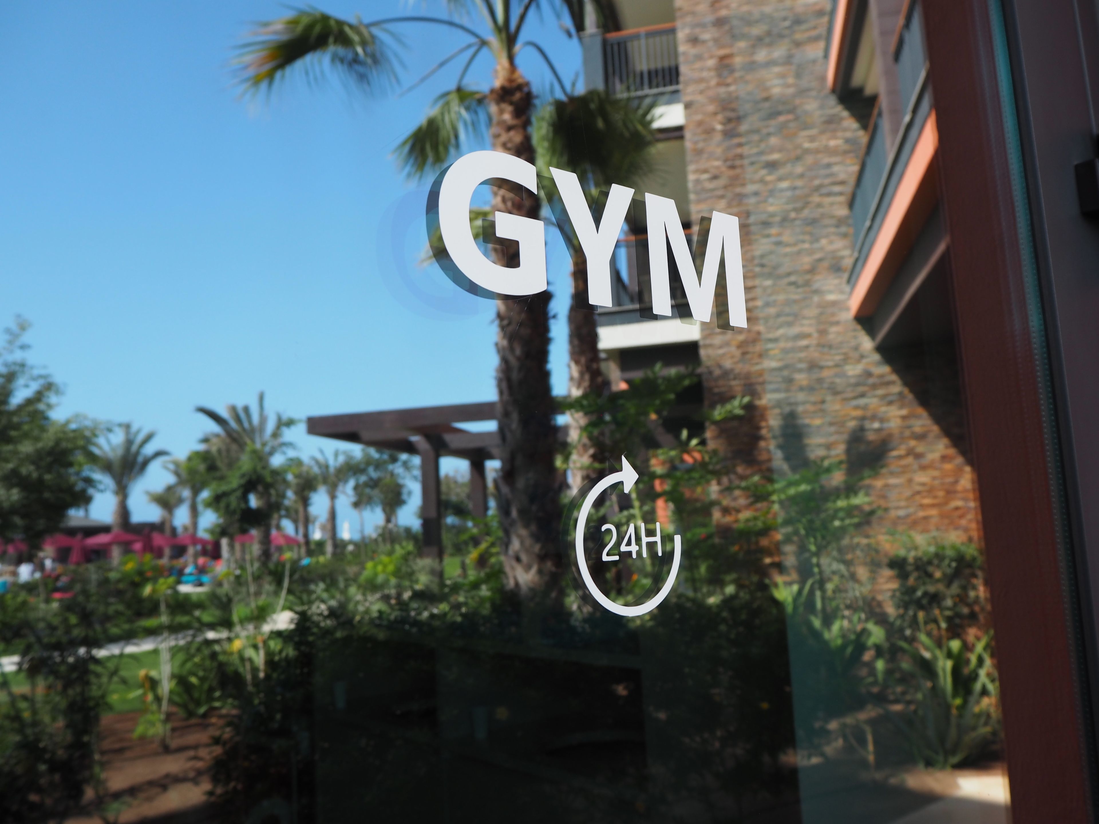 A gym with a 24-hour sign, surrounded by palm trees and a stone building.