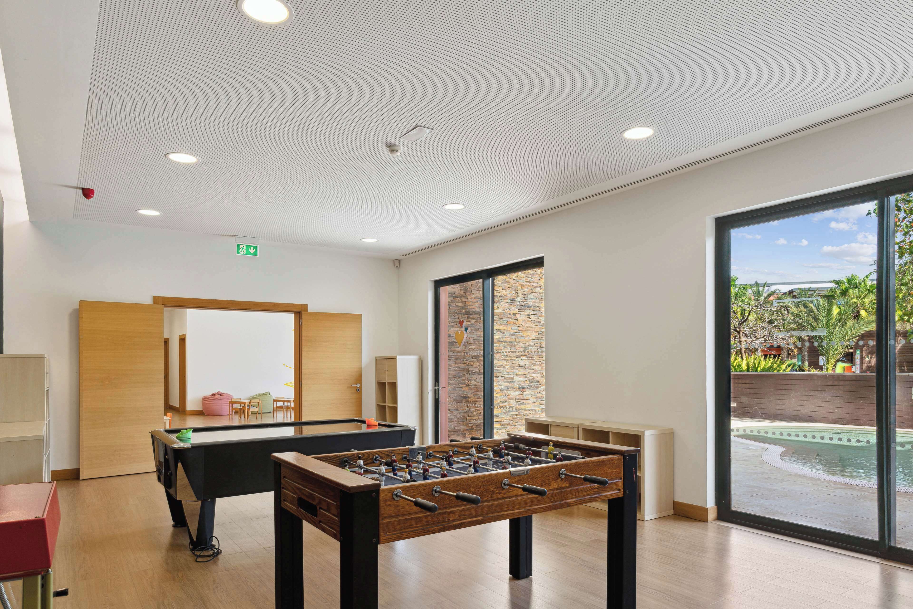 Zona interior de piscina moderna con dos mesas de fútbol de mesa, un banco de madera y vista a una piscina exterior a través de amplios ventanales.