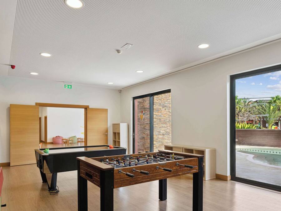 Zona interior de piscina moderna con dos mesas de fútbol de mesa, un banco de madera y vista a una piscina exterior a través de amplios ventanales.