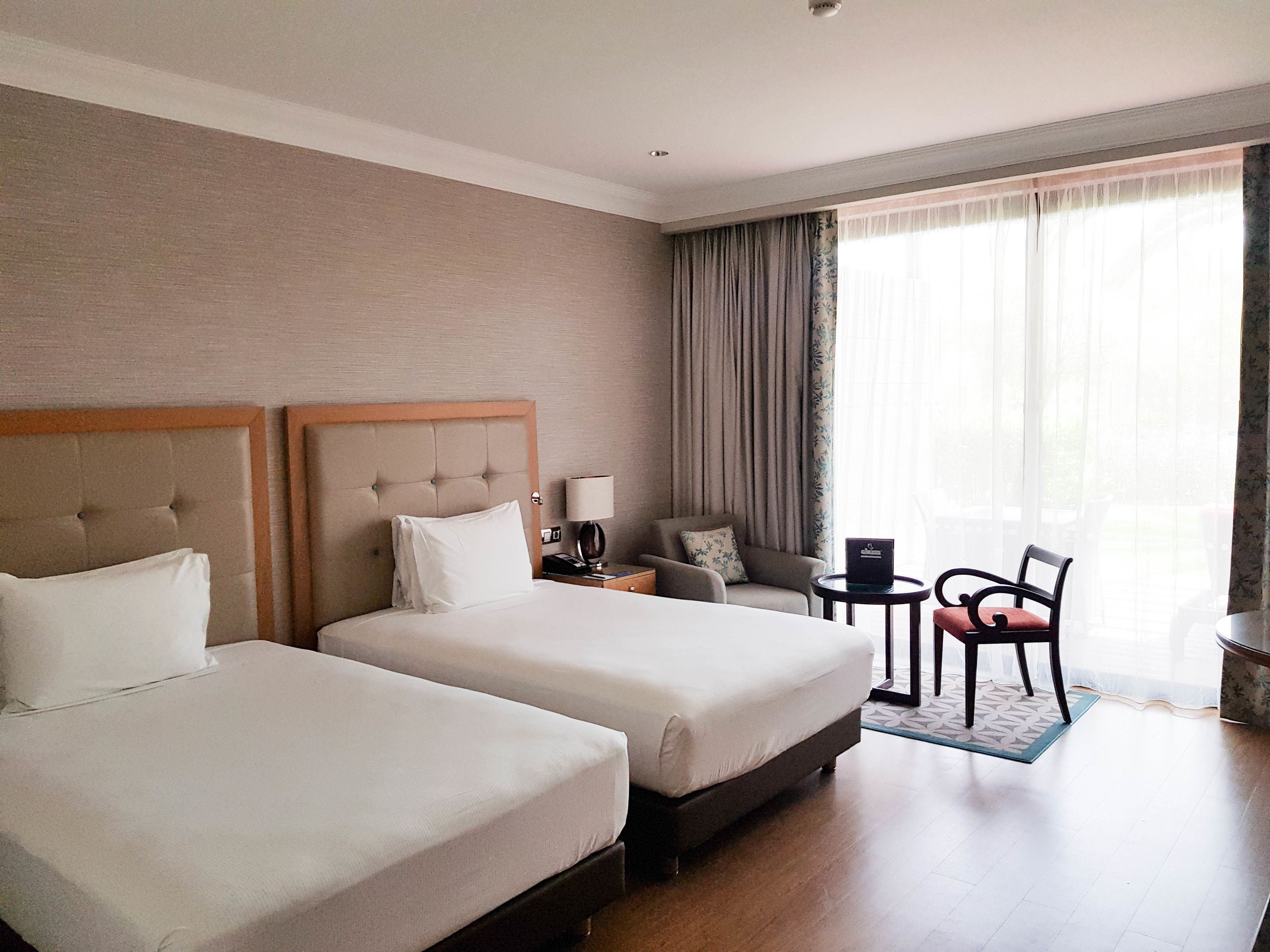 Quarto de hotel com duas camas, uma mesa, uma cadeira e uma janela com cortinas.