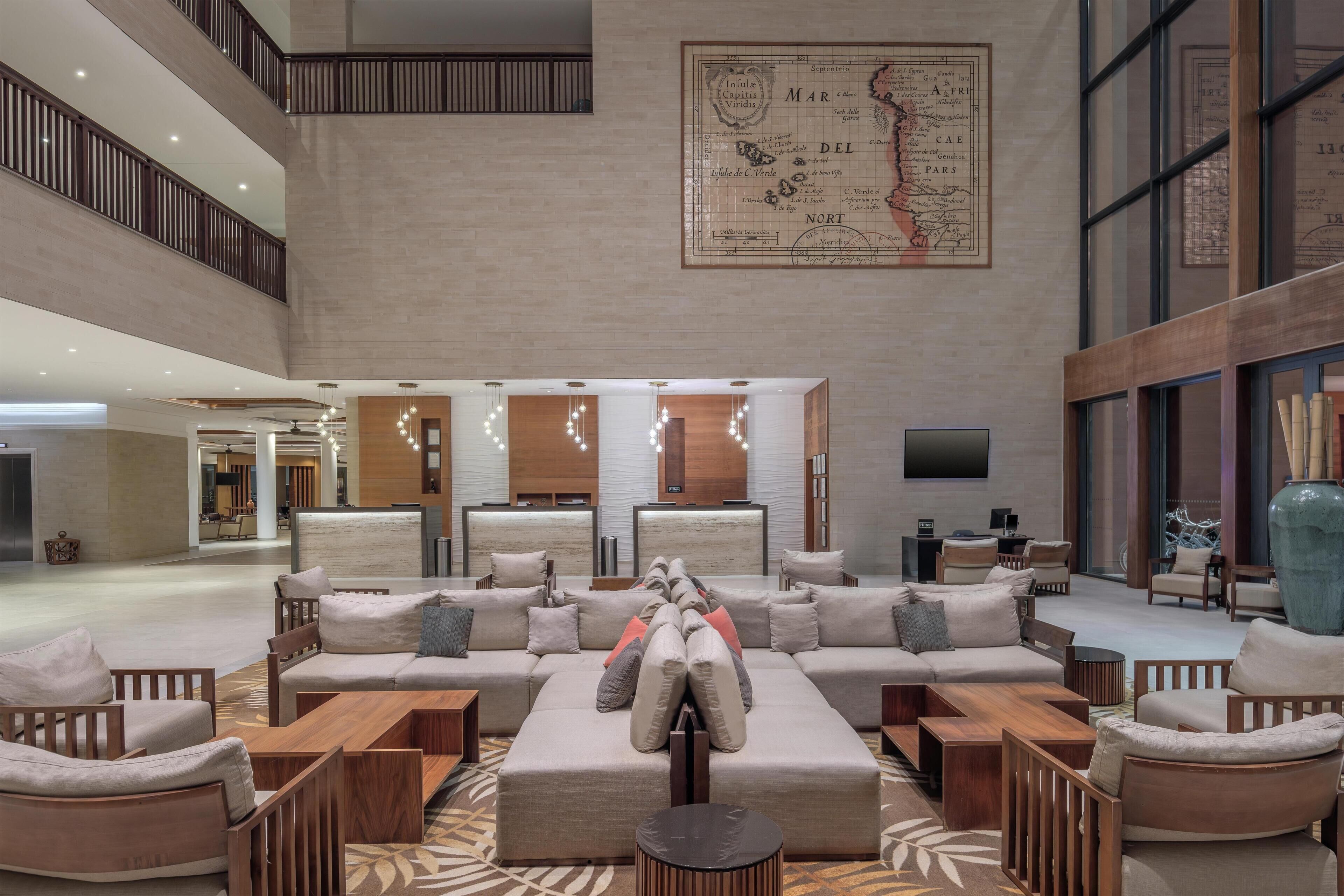 A spacious lobby with a high ceiling, a central seating area with sofas and chairs, a reception desk, and a large map on the wall.