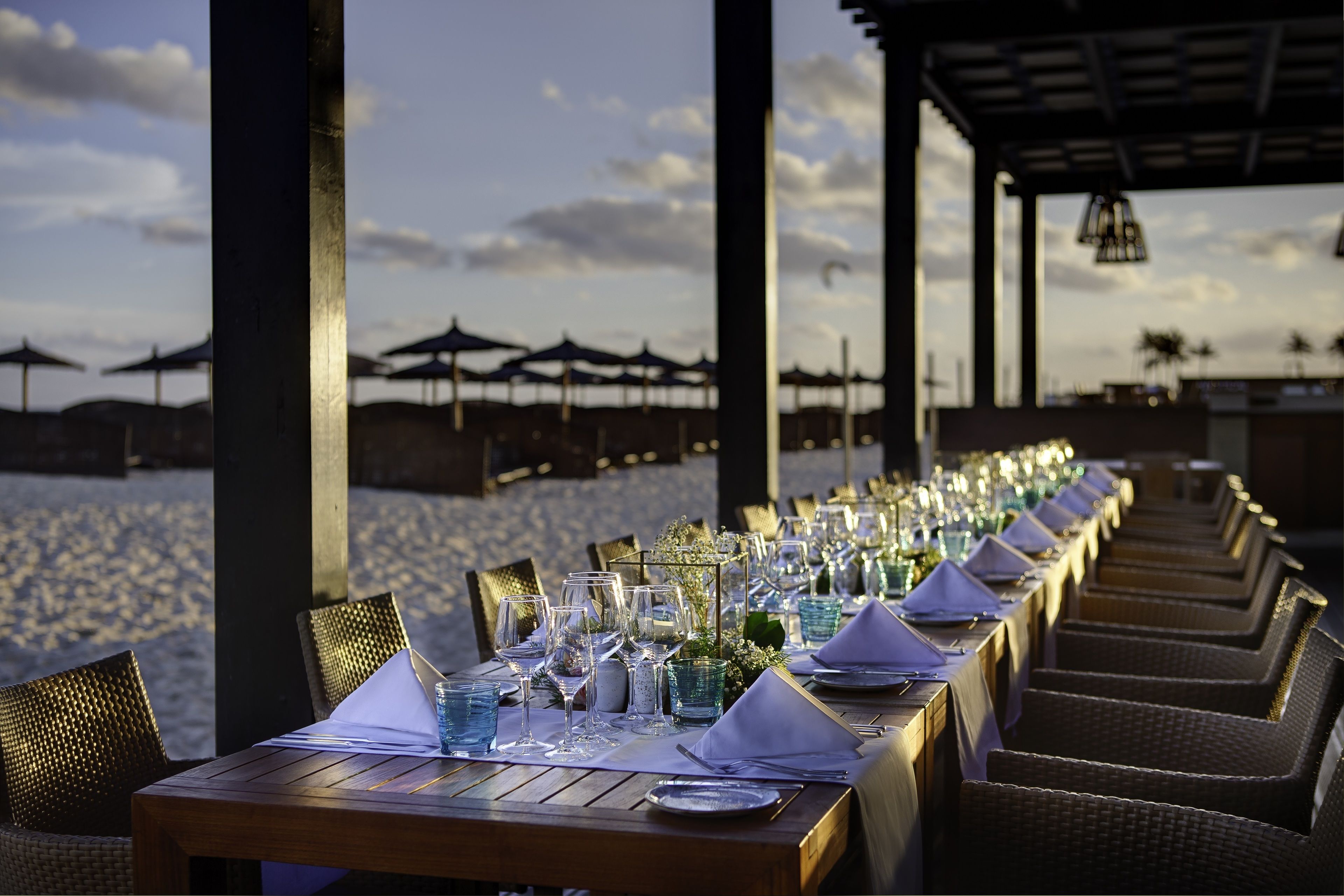 A long dining table set for a meal with chairs on both sides, overlooking a beach with umbrellas in the background.