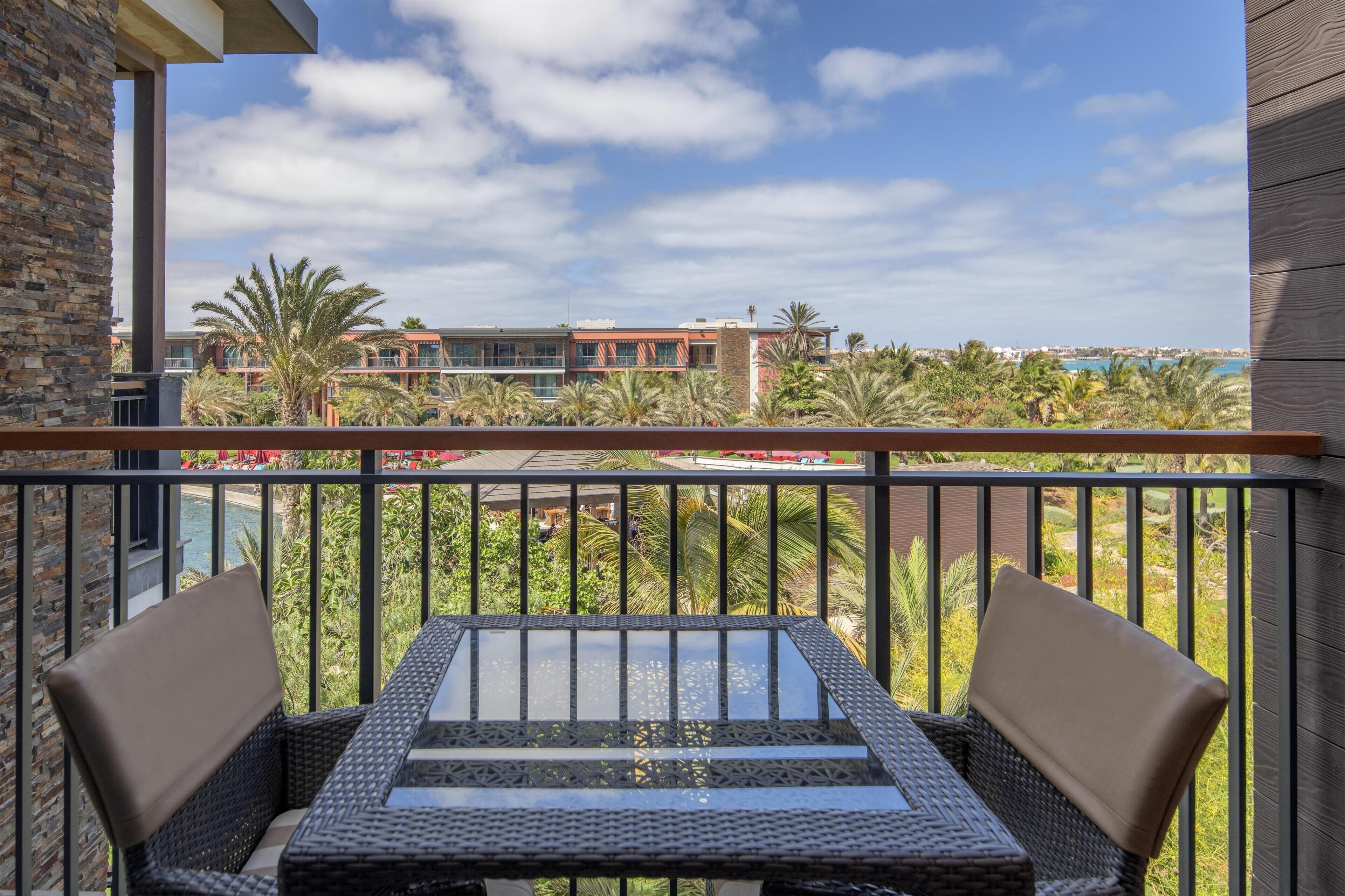 A balcony with wicker furniture, a glass table, and a view of a resort area with palm trees and buildings.