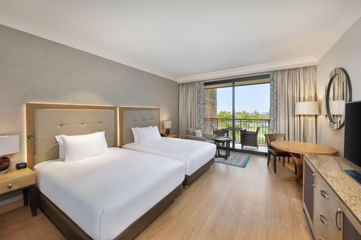 Habitación de hotel con dos camas, un escritorio, una silla, un televisor y un balcón con vistas.