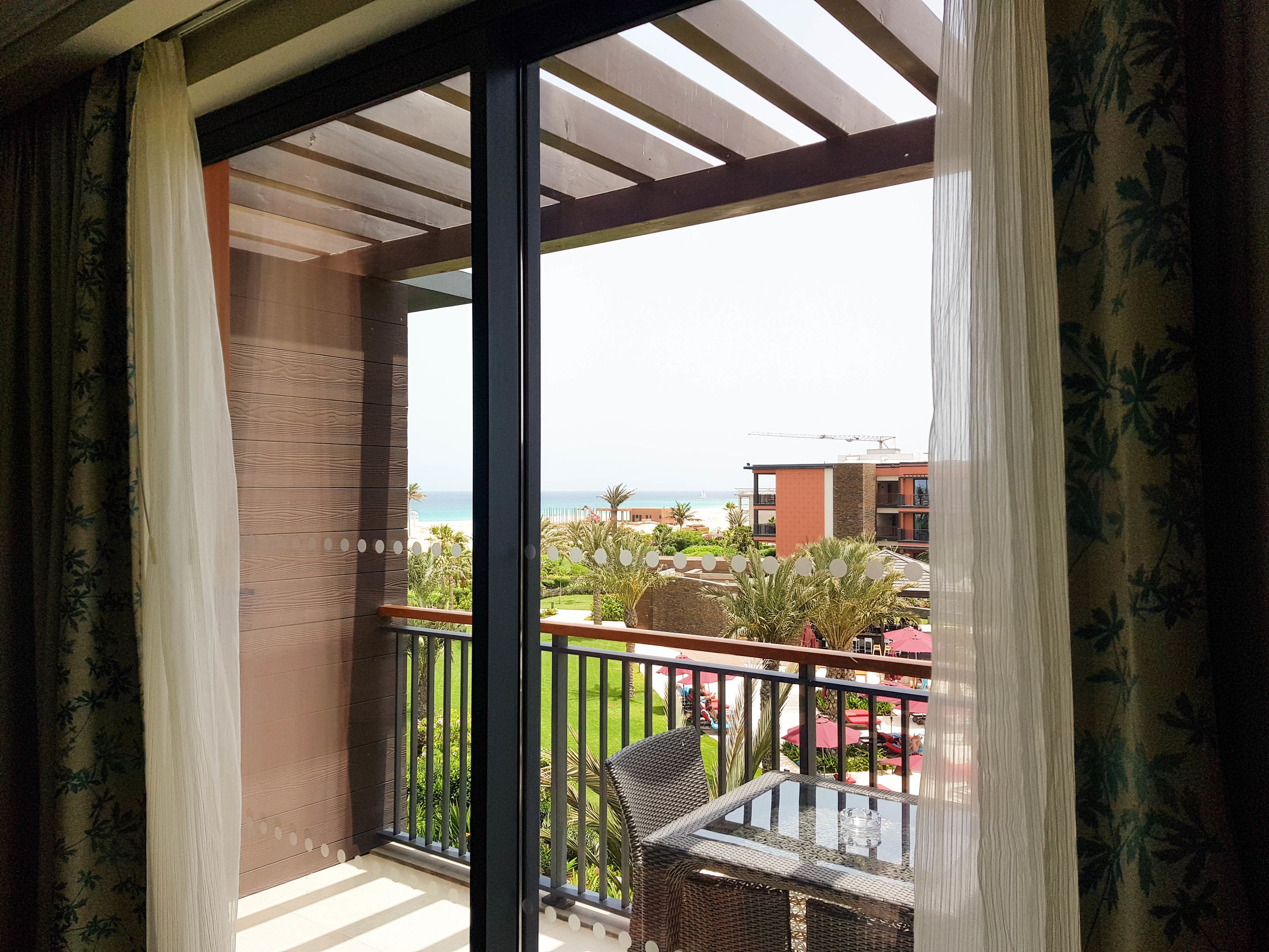 A balcony with a glass table and wicker chairs, overlooking a beach and resort buildings.