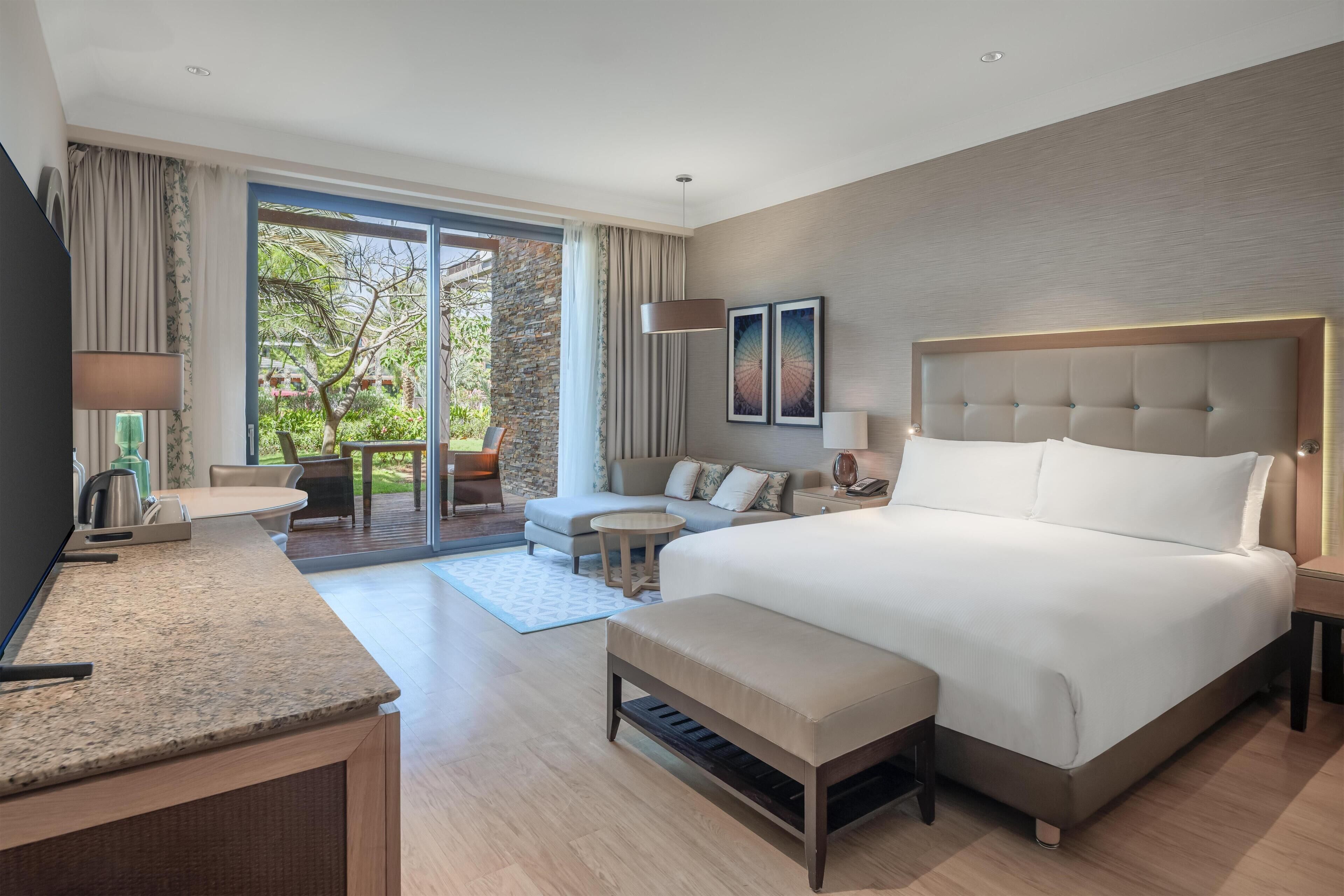 A modern hotel room with a large bed, a desk, a sofa, and a balcony with a view of trees.
