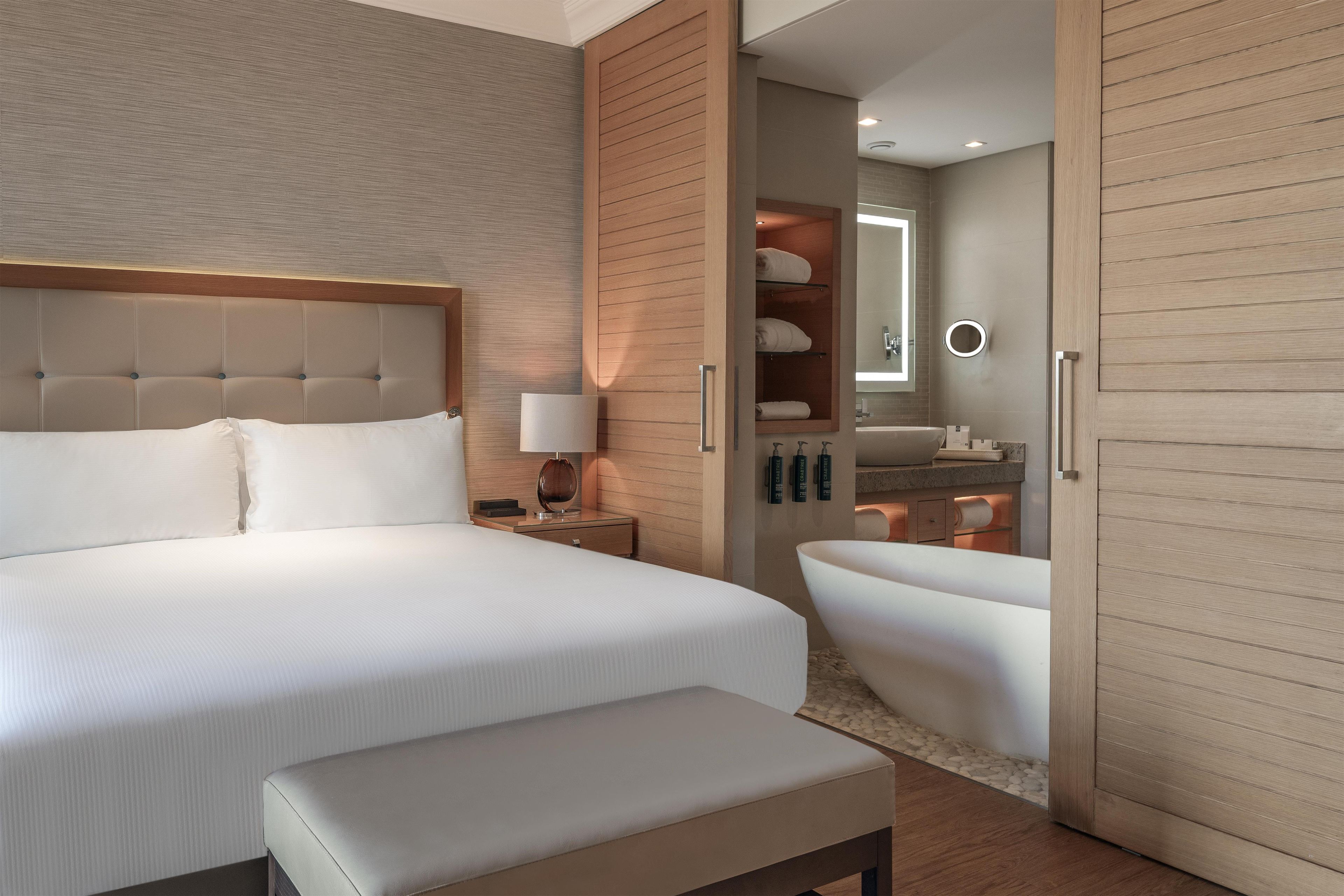 Una habitación de hotel moderna con una cama grande, una mesita de noche con lámpara, un banco integrado, un vestidor y un baño con bañera.