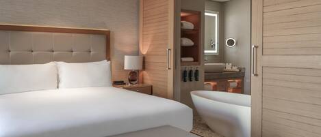 Una habitación de hotel moderna con una cama grande, una mesita de noche con lámpara, un banco integrado, un vestidor y un baño con bañera.