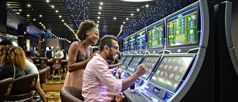 Une zone de jeux de casino avec de nombreux appareils à sous, où l’on voit une femme debout à côté d’un homme qui joue à l’un d’eux.