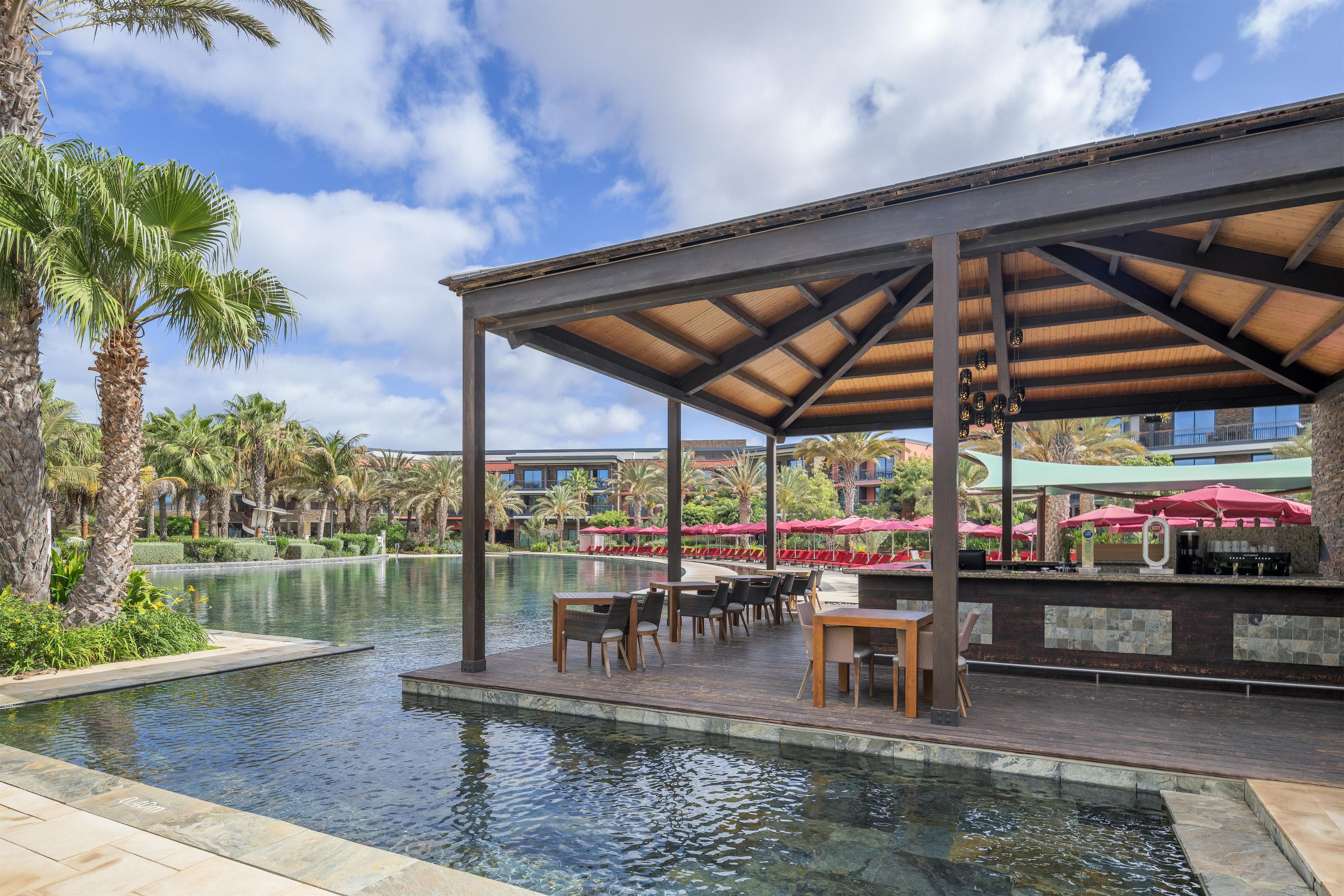 A poolside area with a covered seating area, pool, and palm trees.