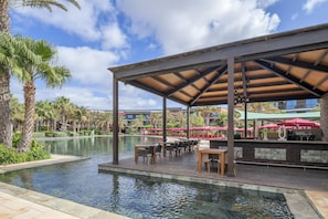 Zona junto a la piscina con un área de descanso cubierta, piscina y palmeras.