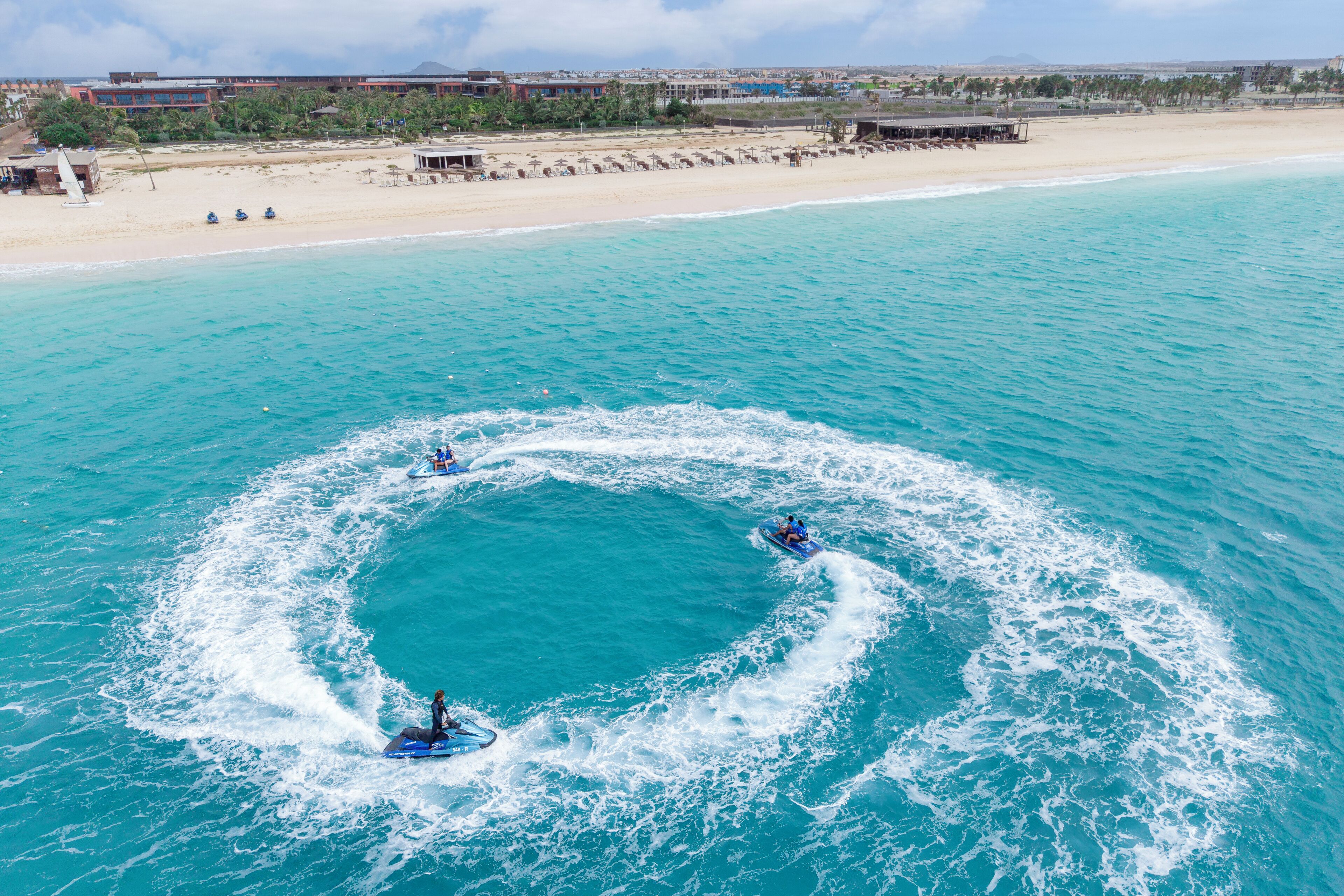 Une plage avec une mer d’un bleu limpide, un rivage sablonneux et un bâtiment en arrière-plan.
