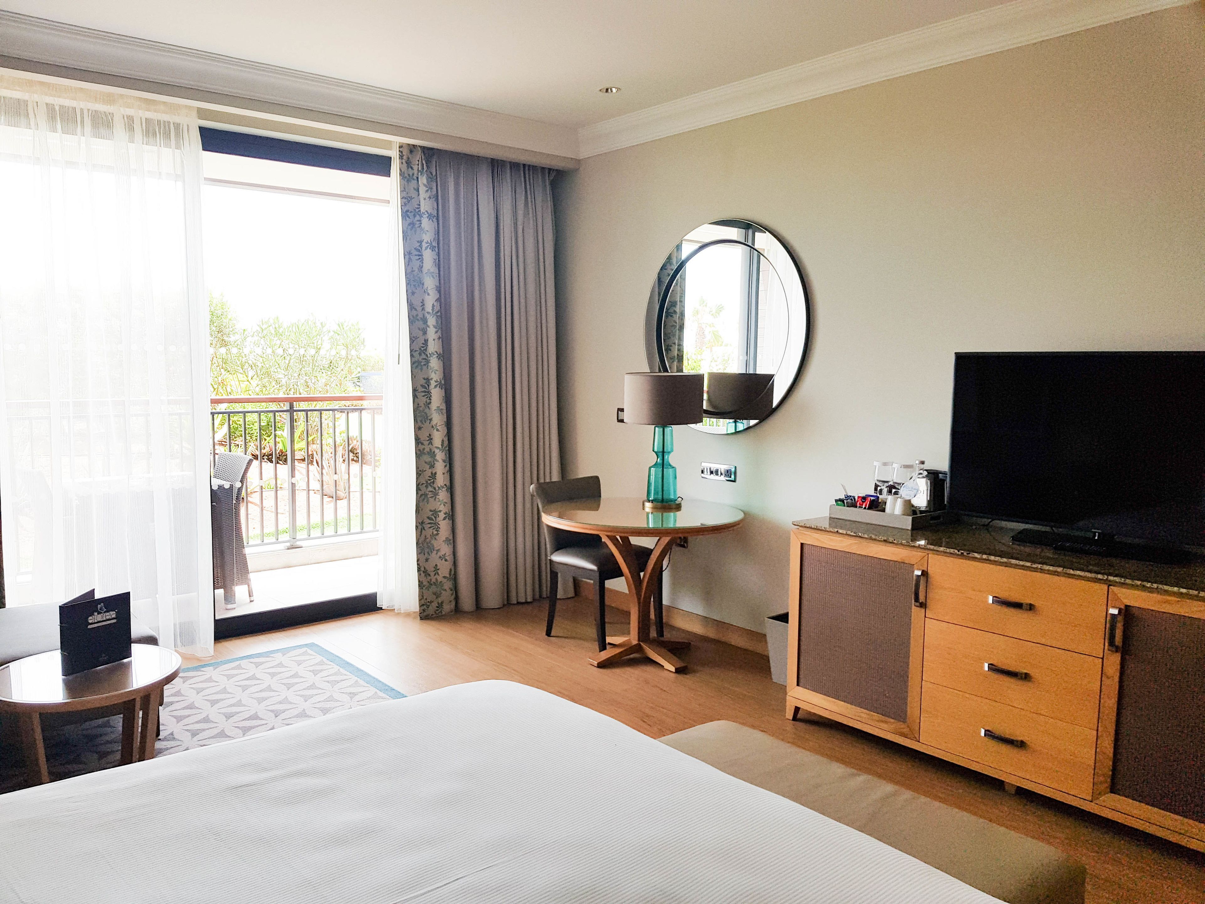 A hotel room with a bed, a desk with a lamp, a television, and a view of the outdoors.