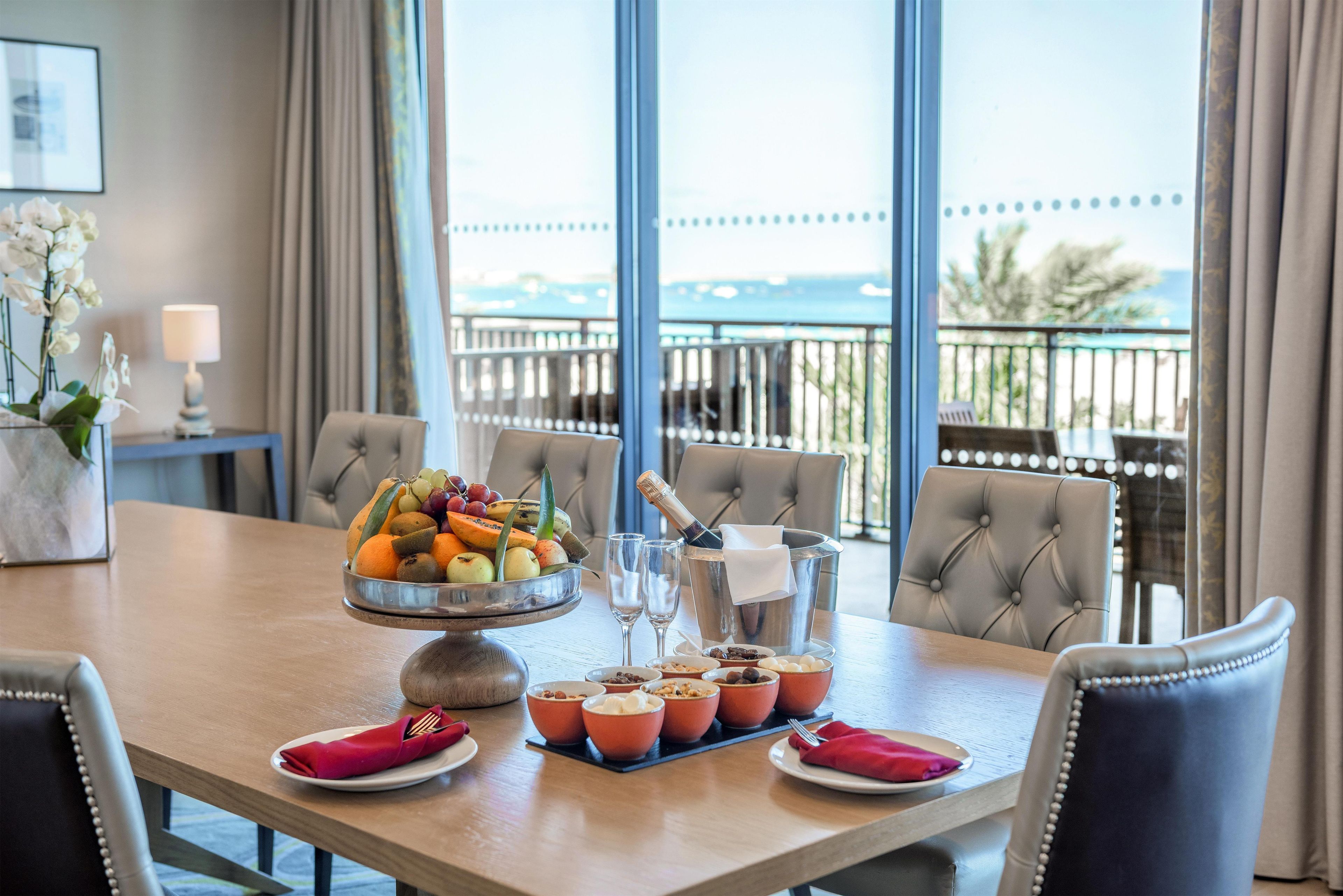 Una mesa de comedor con fruta, una botella de champán y copas, en una habitación con vistas al mar.