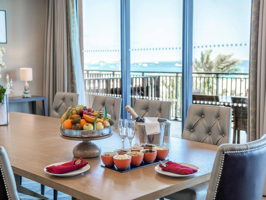 Una mesa de comedor con fruta, una botella de champán y copas, en una habitación con vistas al mar.