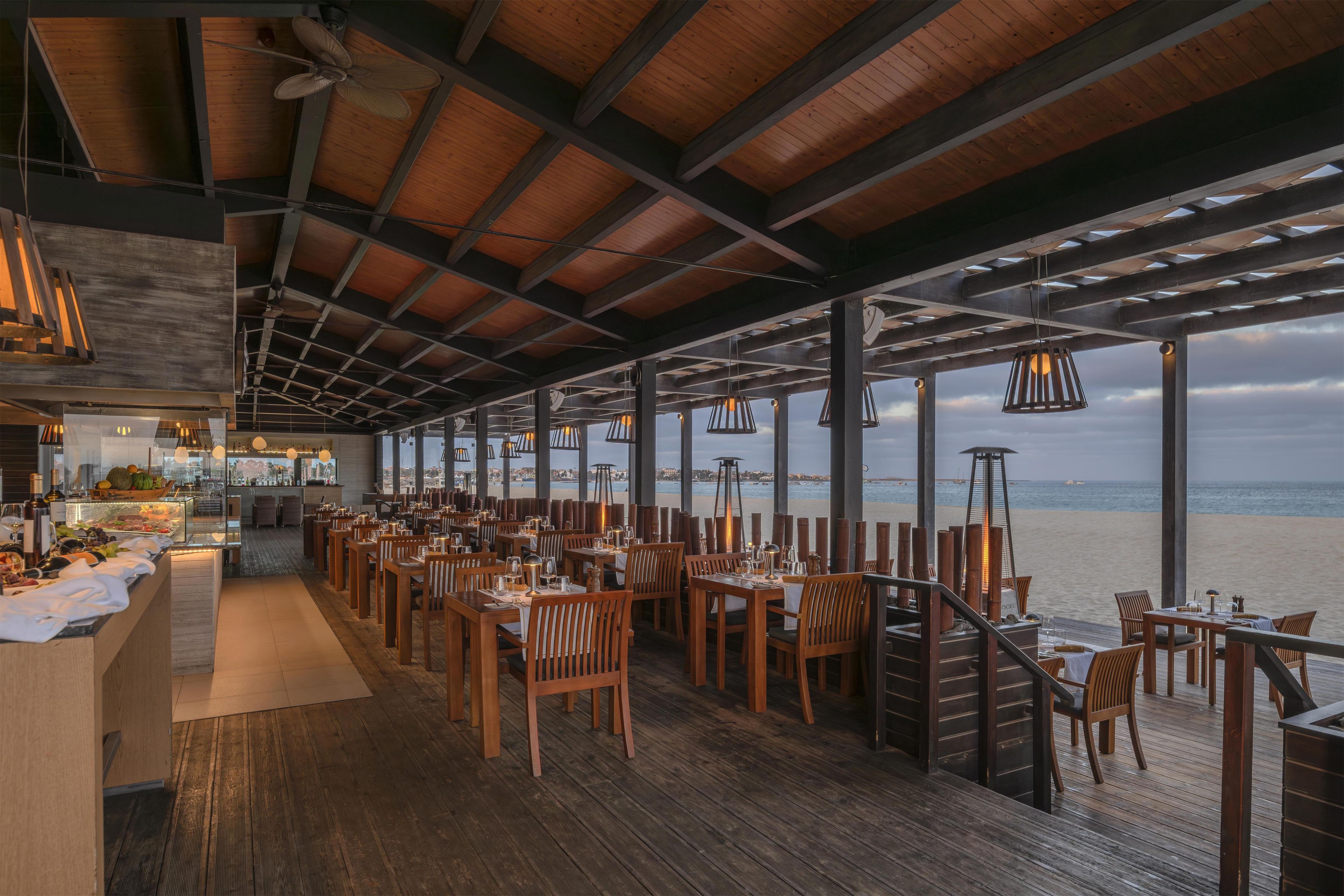 A restaurant with wooden flooring, tables, chairs, and a view of the sea.