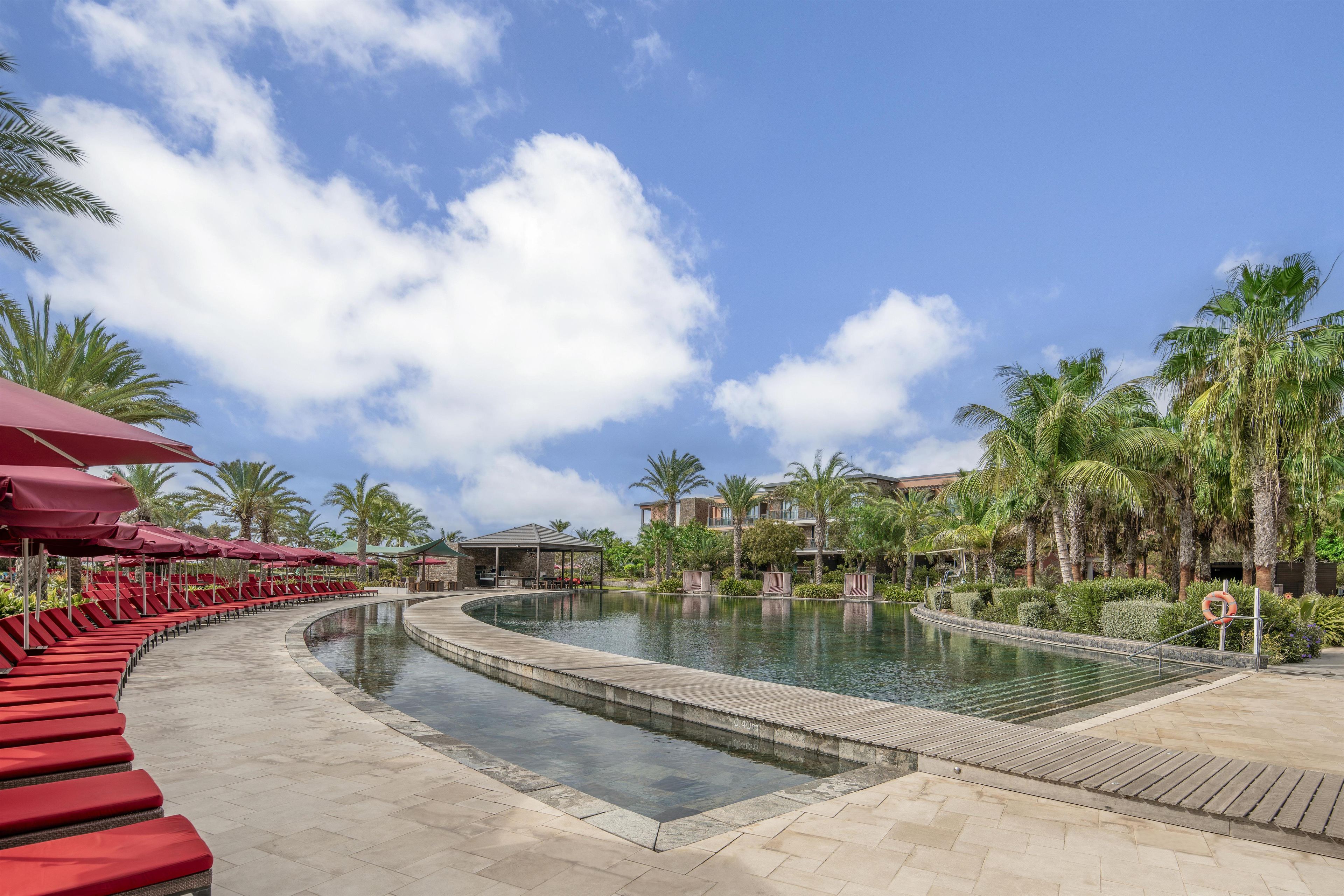 Zona de piscina en un resort con sillas de descanso rojas, piscina con forma curva y palmeras.