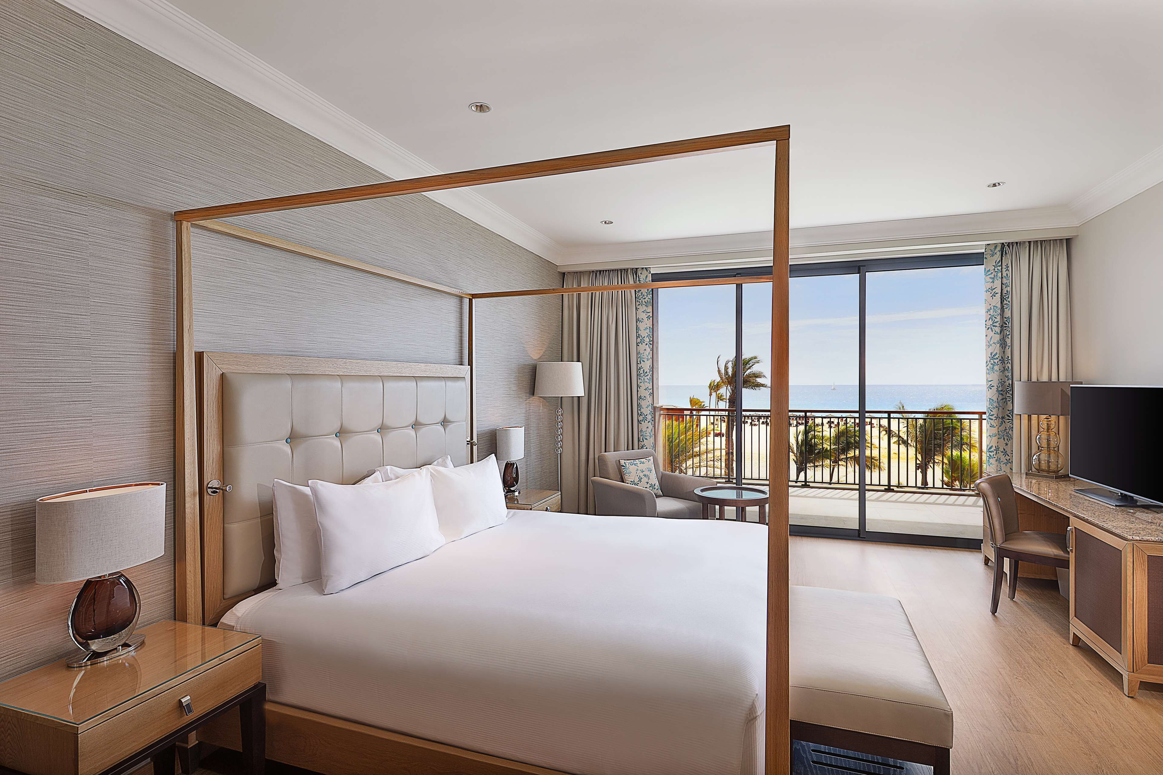 Une chambre d’hôtel avec un lit à baldaquin, un bureau avec une télévision et un balcon offrant une vue sur l’océan.