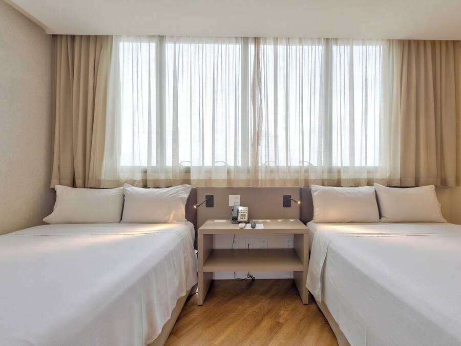 A hotel room with two single beds, a bedside table with a phone and a lamp, and a window with sheer curtains.