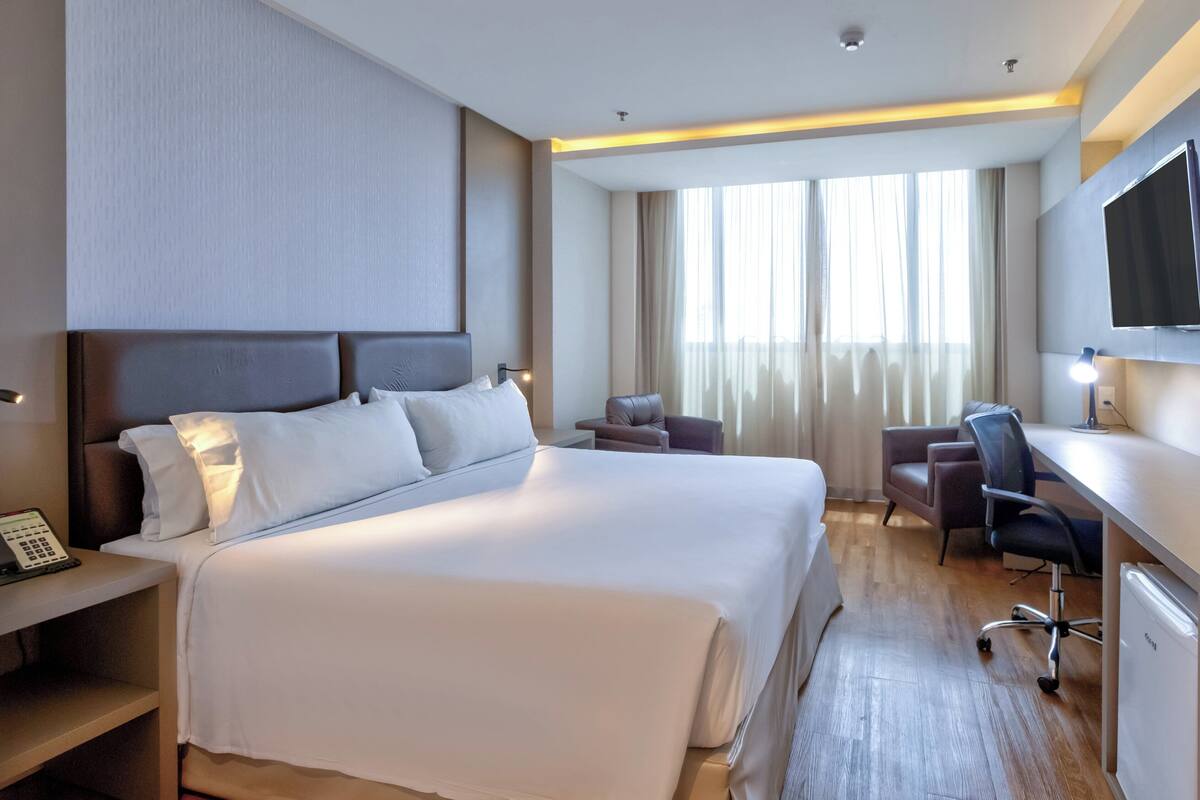 A hotel room with a large bed, a desk with a chair, a television, and a desk lamp.