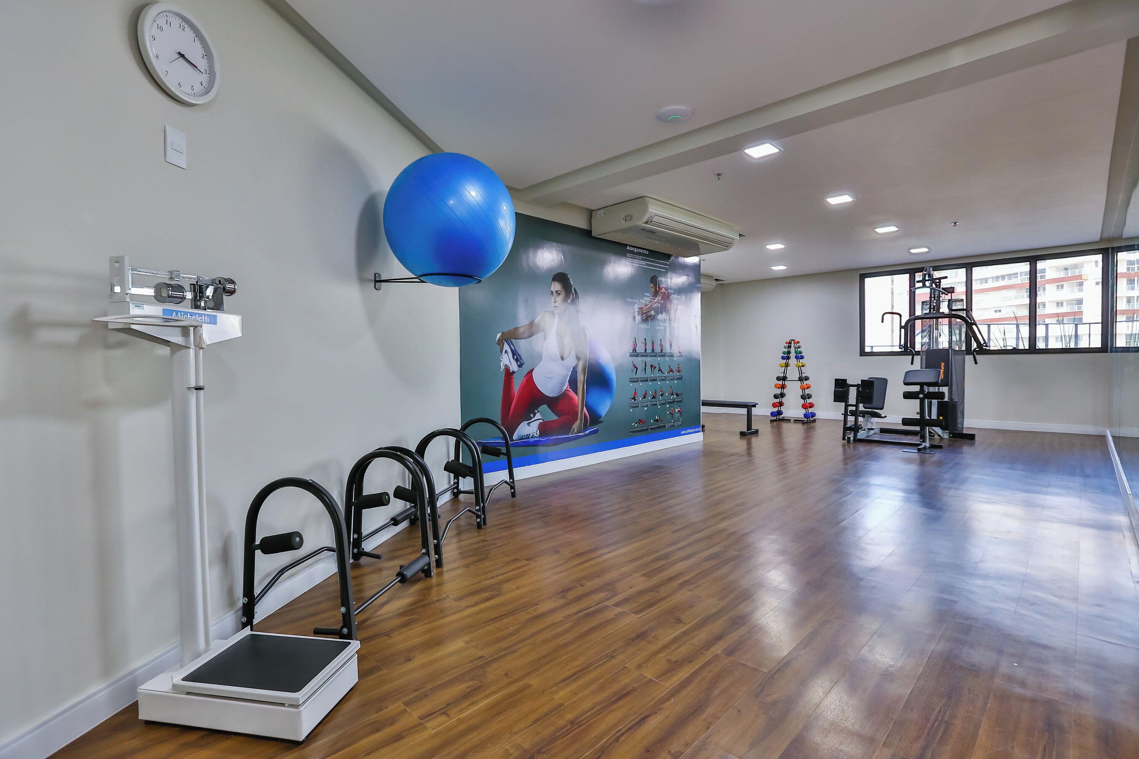 Uma academia espaçosa com equipamentos de ginástica, uma bola de exercício azul grande e um painel com pôsteres de fitness.