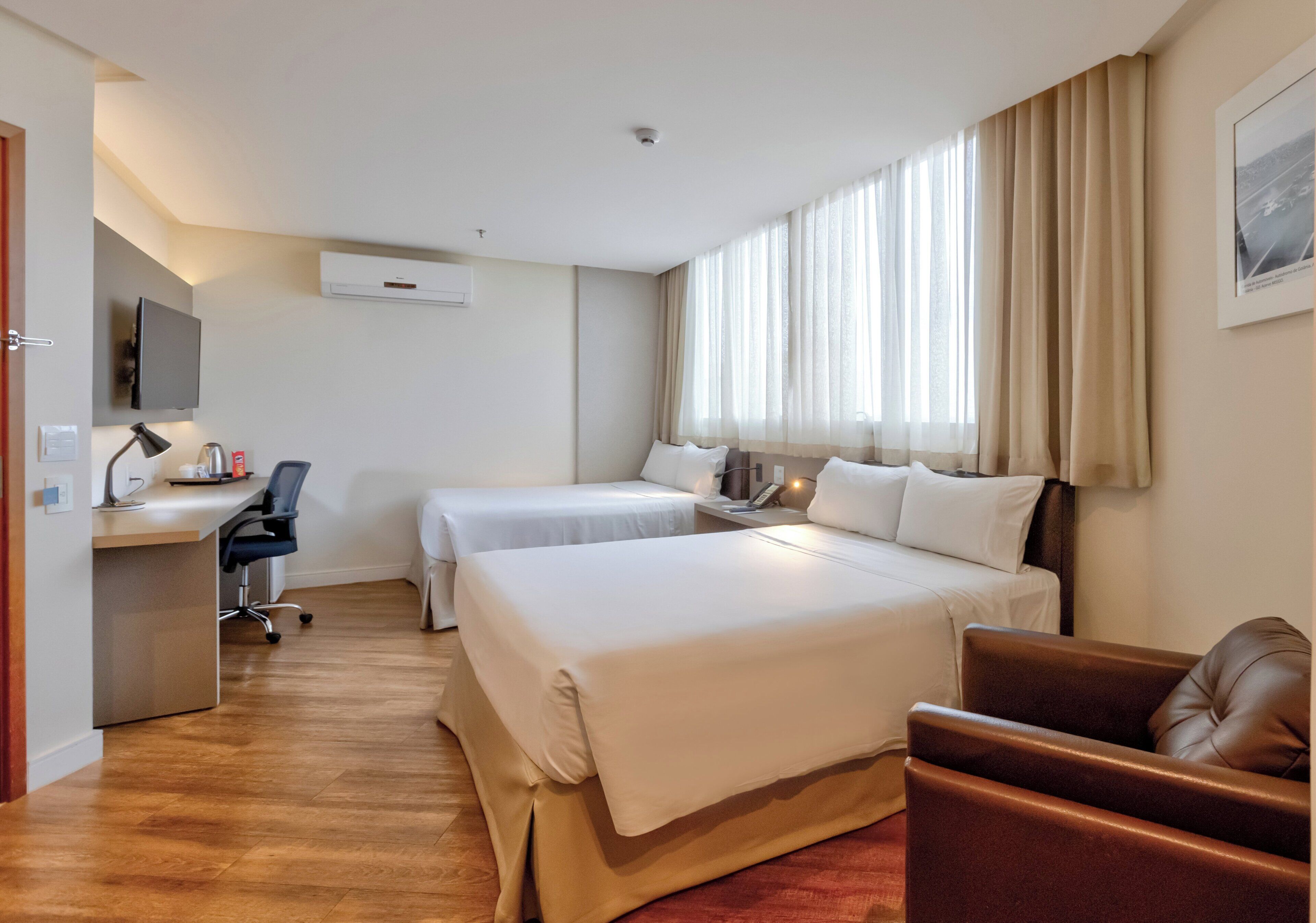 Quarto de hotel com duas camas, uma escrivaninha com cadeira, uma televisão e uma janela com cortinas.