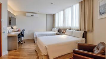 Quarto de hotel com duas camas, uma escrivaninha com cadeira, uma televisão e uma janela com cortinas.