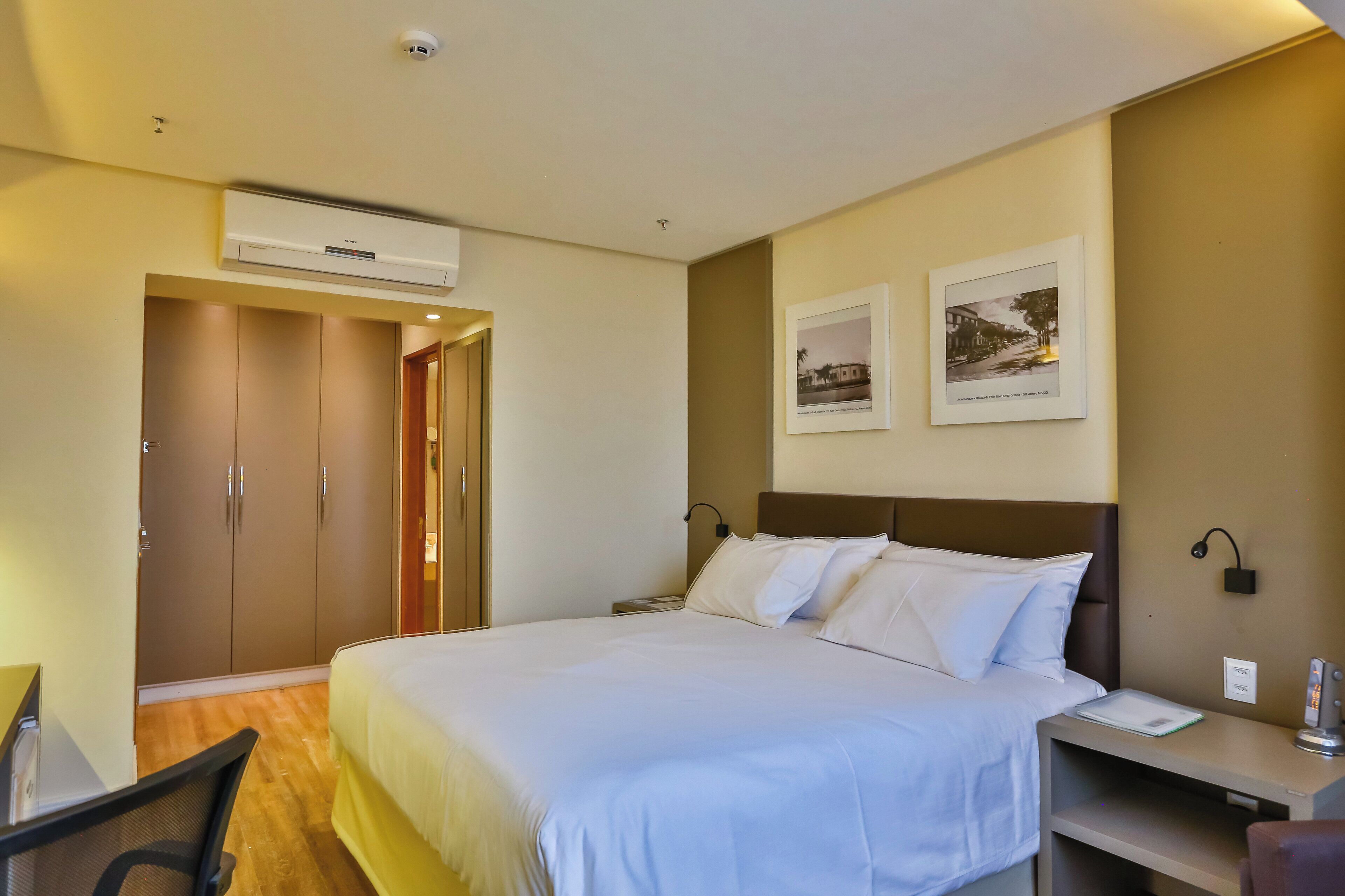 A hotel room with a bed, a desk, a chair, a wardrobe, and two framed pictures on the wall.