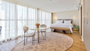 Une chambre d’hôtel moderne avec un grand lit, une table ronde avec des chaises, une petite table d’appoint et une grande fenêtre avec des rideaux légers.