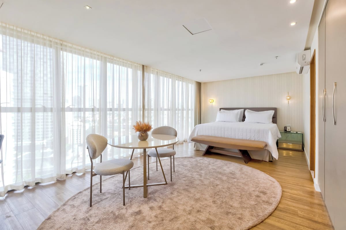 A modern hotel room with a large bed, a round table with chairs, a small side table, and a large window with sheer curtains.
