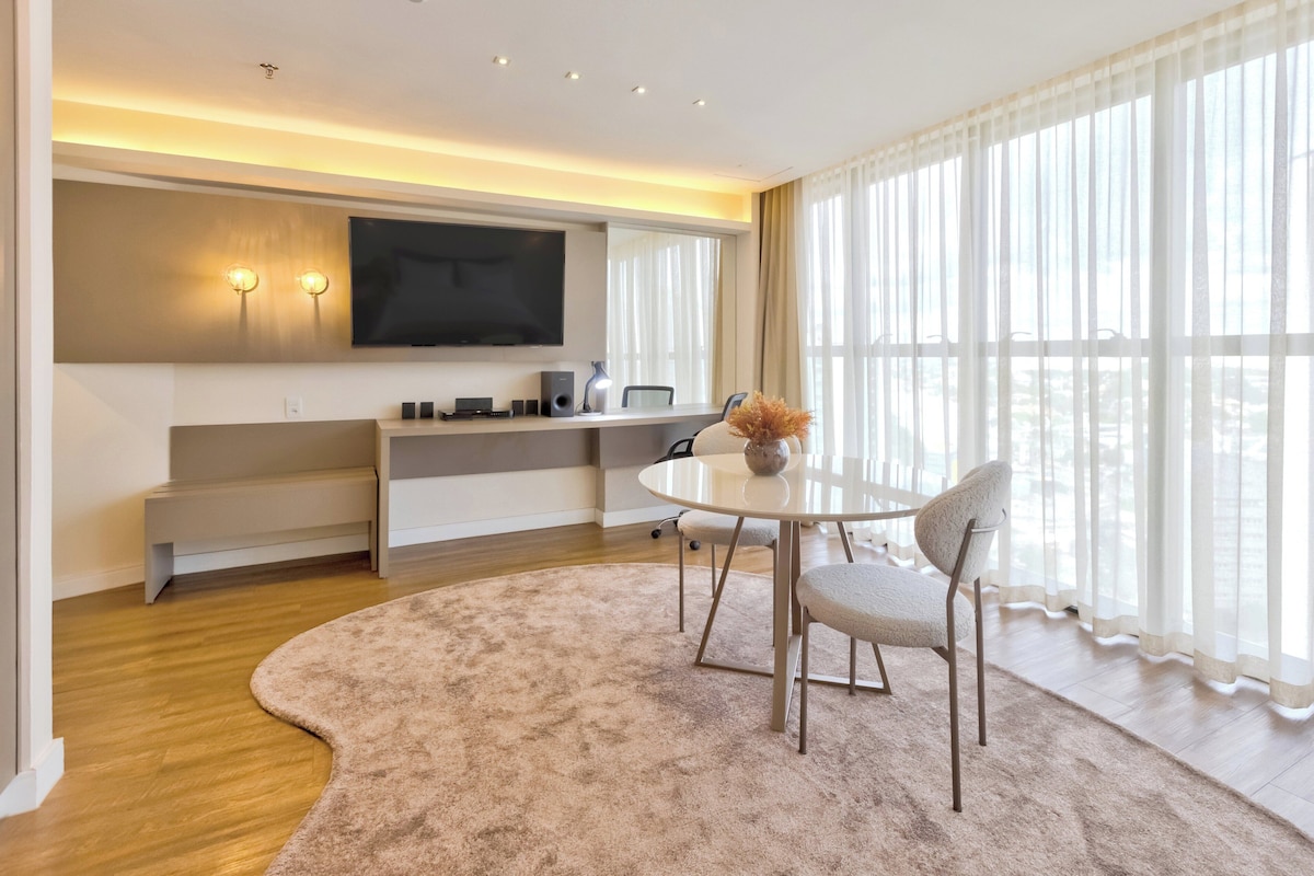 A modern living room with a flat-screen TV, a round table, and a large window with sheer curtains.
