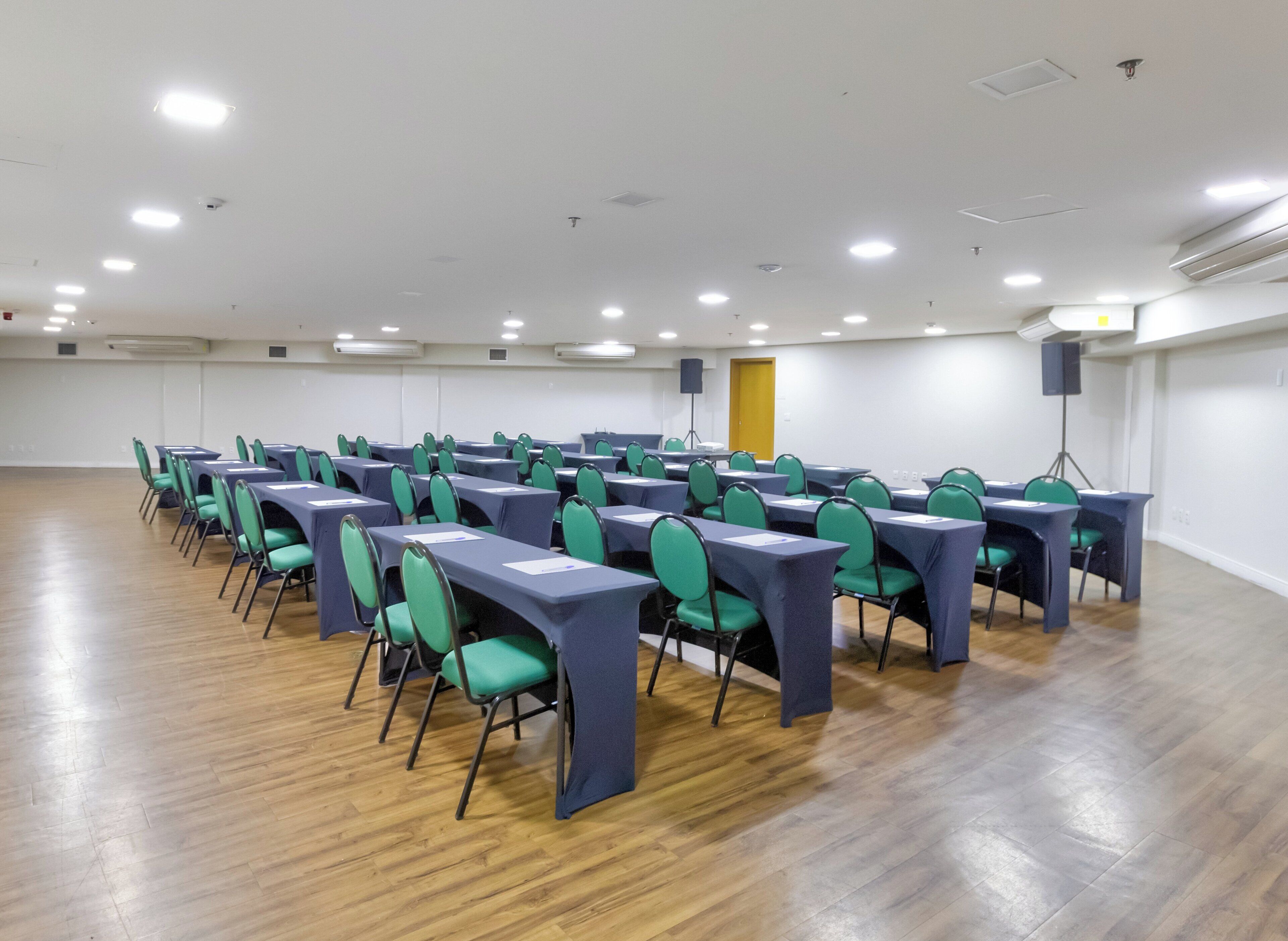 Un salón de conferencias con filas de mesas y sillas, paredes blancas y suelo de madera.
