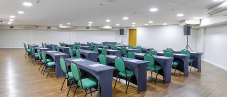 Un salón de conferencias con filas de mesas y sillas, paredes blancas y suelo de madera.