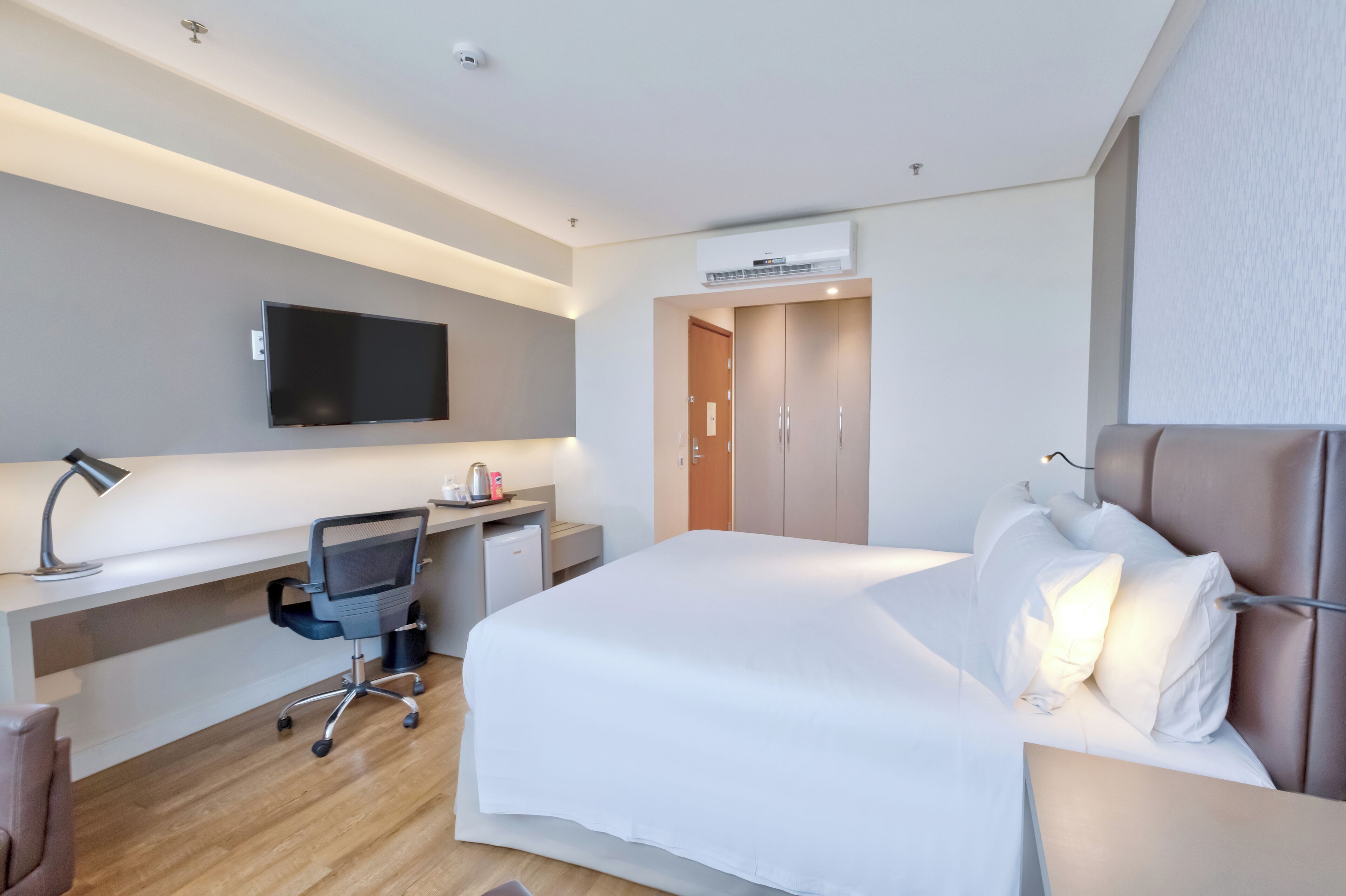 Quarto de hotel com cama, escrivaninha, cadeira, televisão e guarda-roupa.