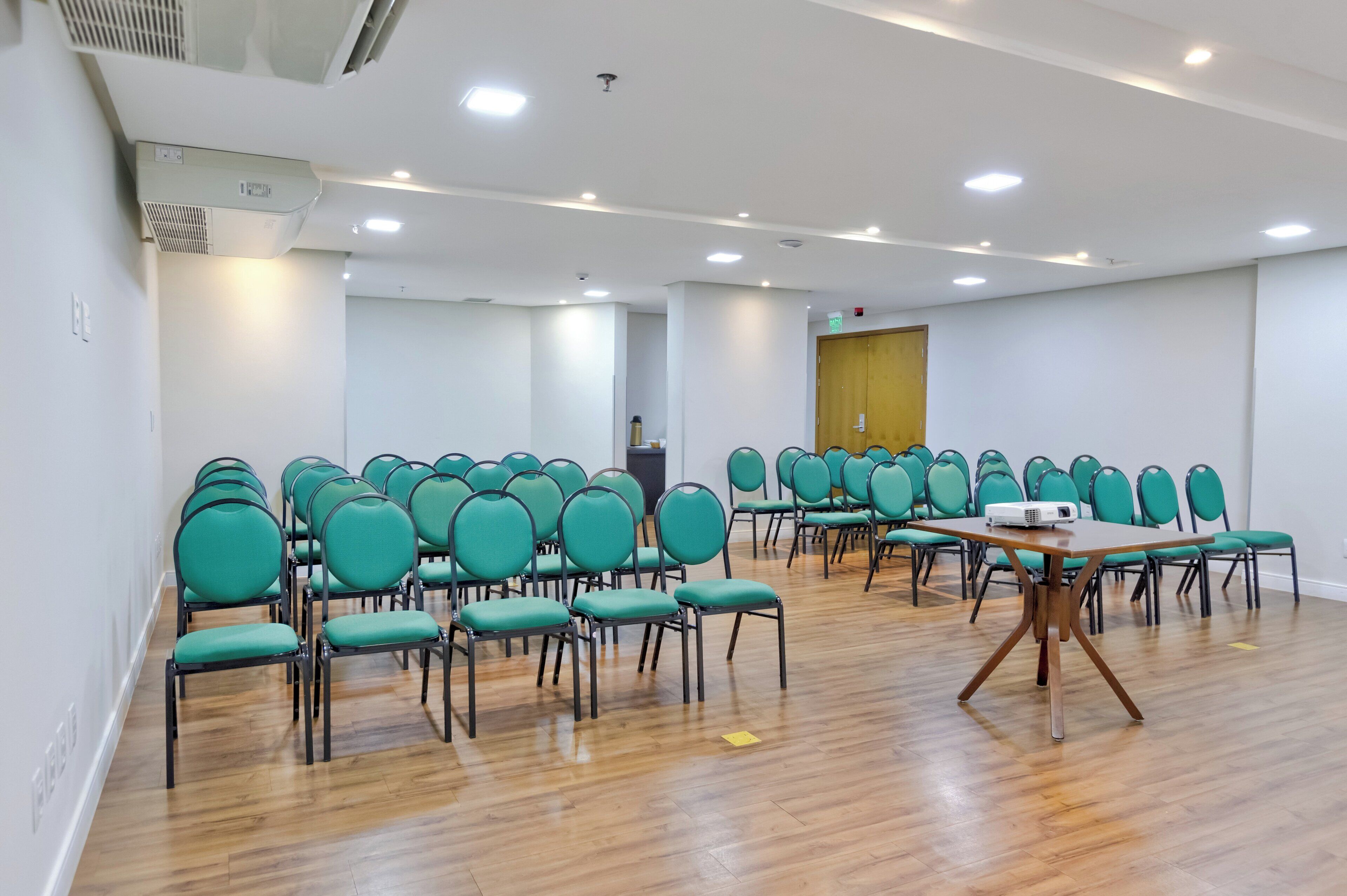 A conference room with rows of green chairs, a wooden table with a projector, and a wooden door in the background.