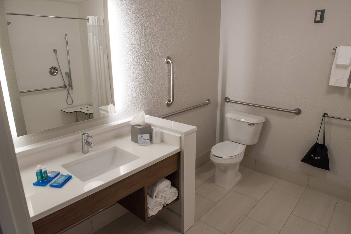 A modern bathroom with a white sink, mirror, and toilet. There are handrails on the walls and a towel rack.