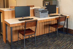 Quarto de hotel com escrivaninha de madeira, dois monitores de computador, uma impressora e duas cadeiras.