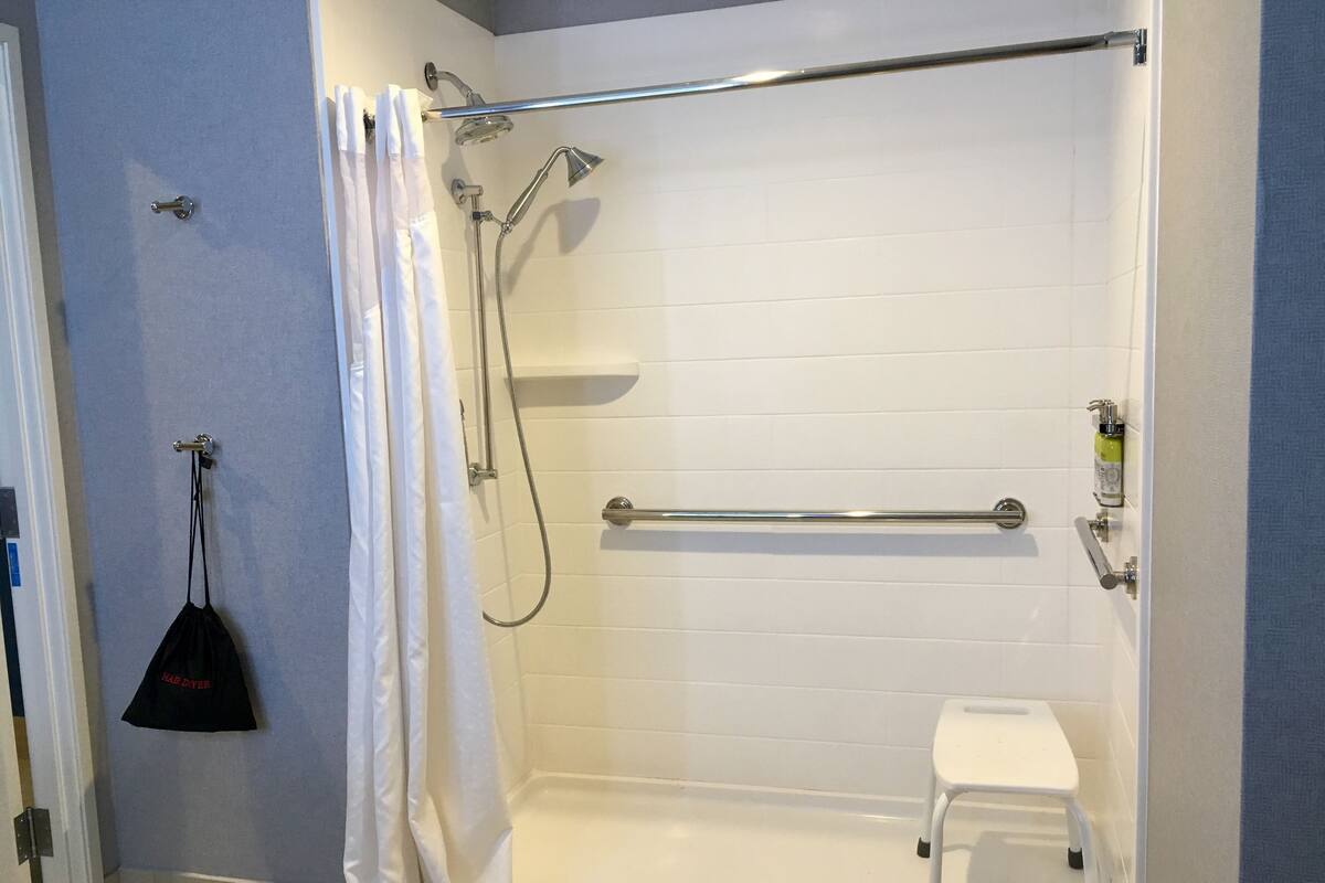 Une douche à l’italienne avec un mur recouvert de carreaux blancs, un pommeau de douche, un flexible de douche, un porte-serviettes et un petit tabouret blanc.