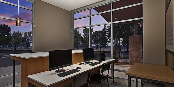 A modern office space with two desks, computers, and a view of trees and sky through large windows.