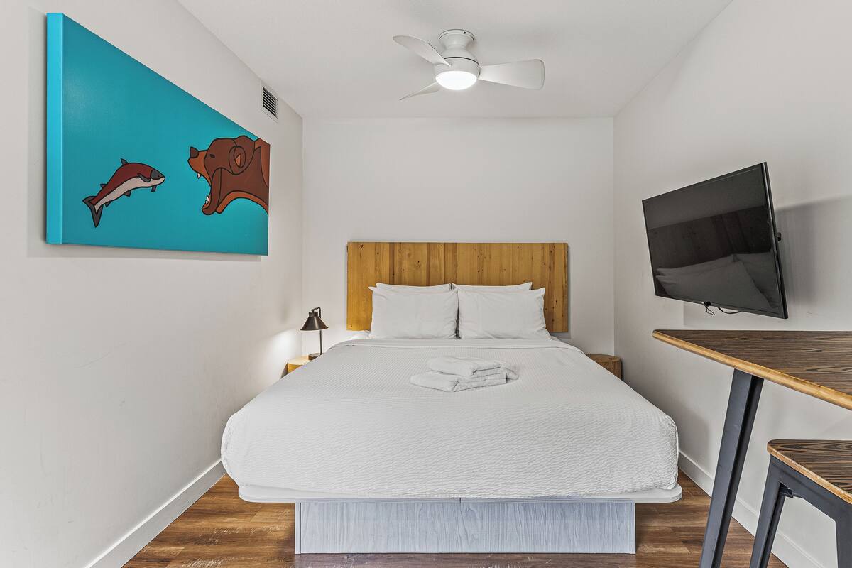 A bedroom with a bed, a ceiling fan, a TV, and a painting on the wall.