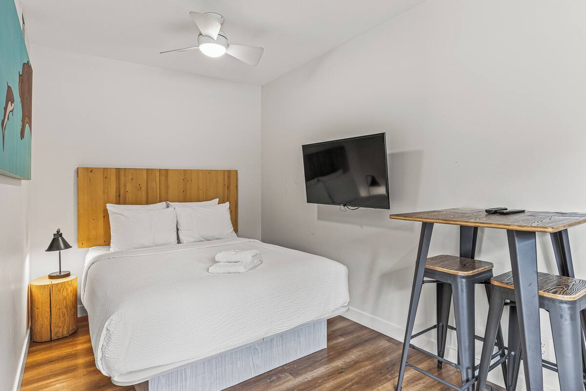 A bedroom with a bed, a wooden headboard, a ceiling fan, a TV mounted on the wall, and a small table with two stools.