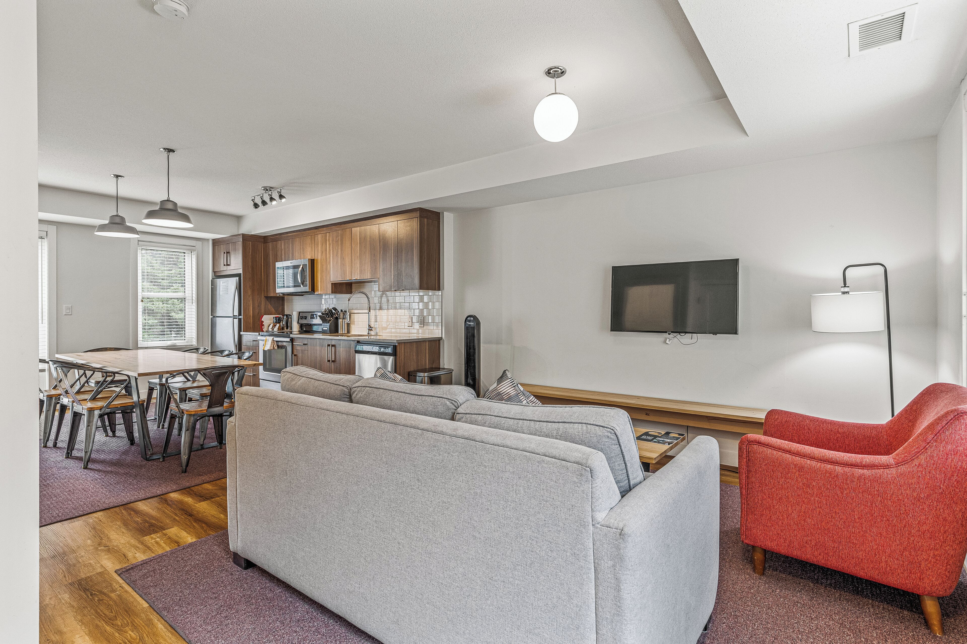 Una sala moderna con un sofá gris, un sillón rojo y un comedor con una mesa y sillas de madera.
