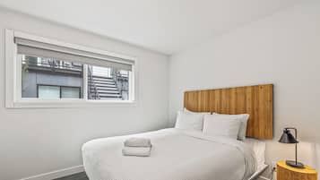 A neatly made bed with a white comforter and pillows, a wooden headboard, a bedside table with a lamp, and a window with blinds.