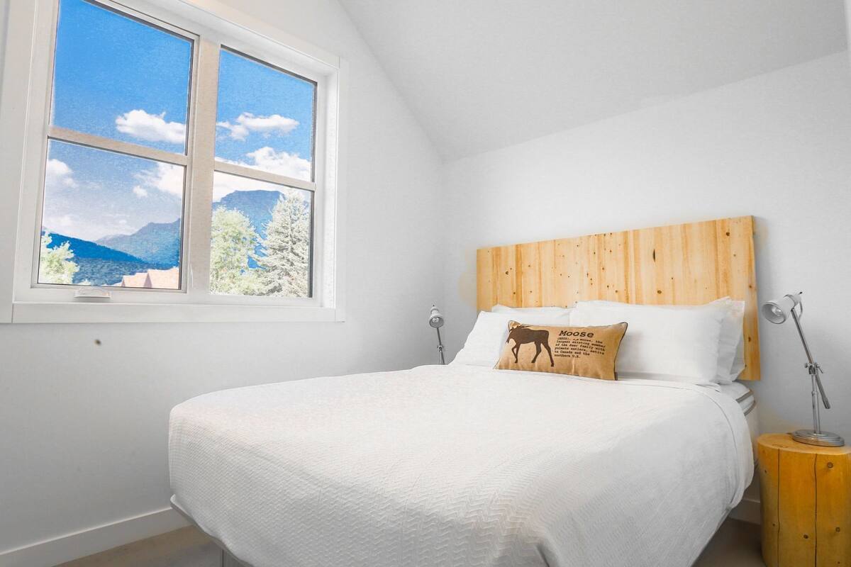 A bedroom with a bed, a wooden headboard, a window with a view of mountains and trees, and a bedside lamp.