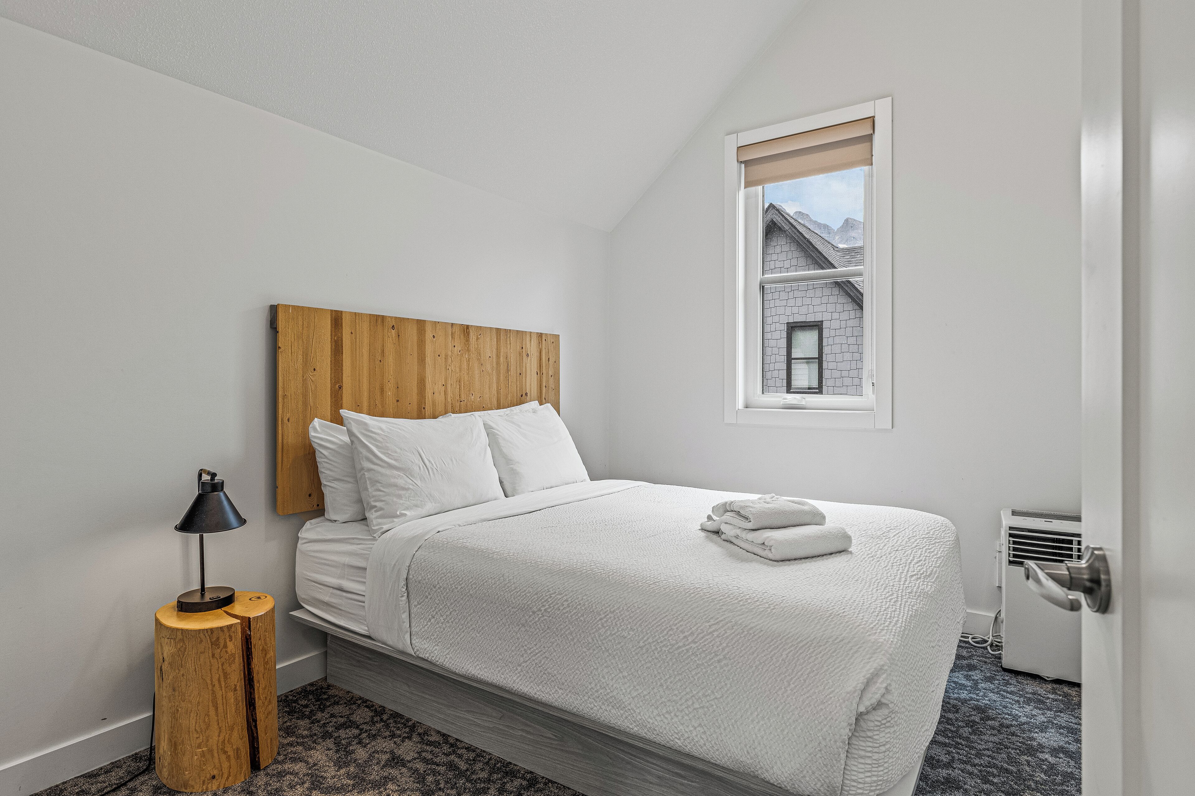 A neatly made bed with white bedding, a wooden headboard, and a bedside lamp on a round wooden nightstand. A window with a view of a building is visible in the background.